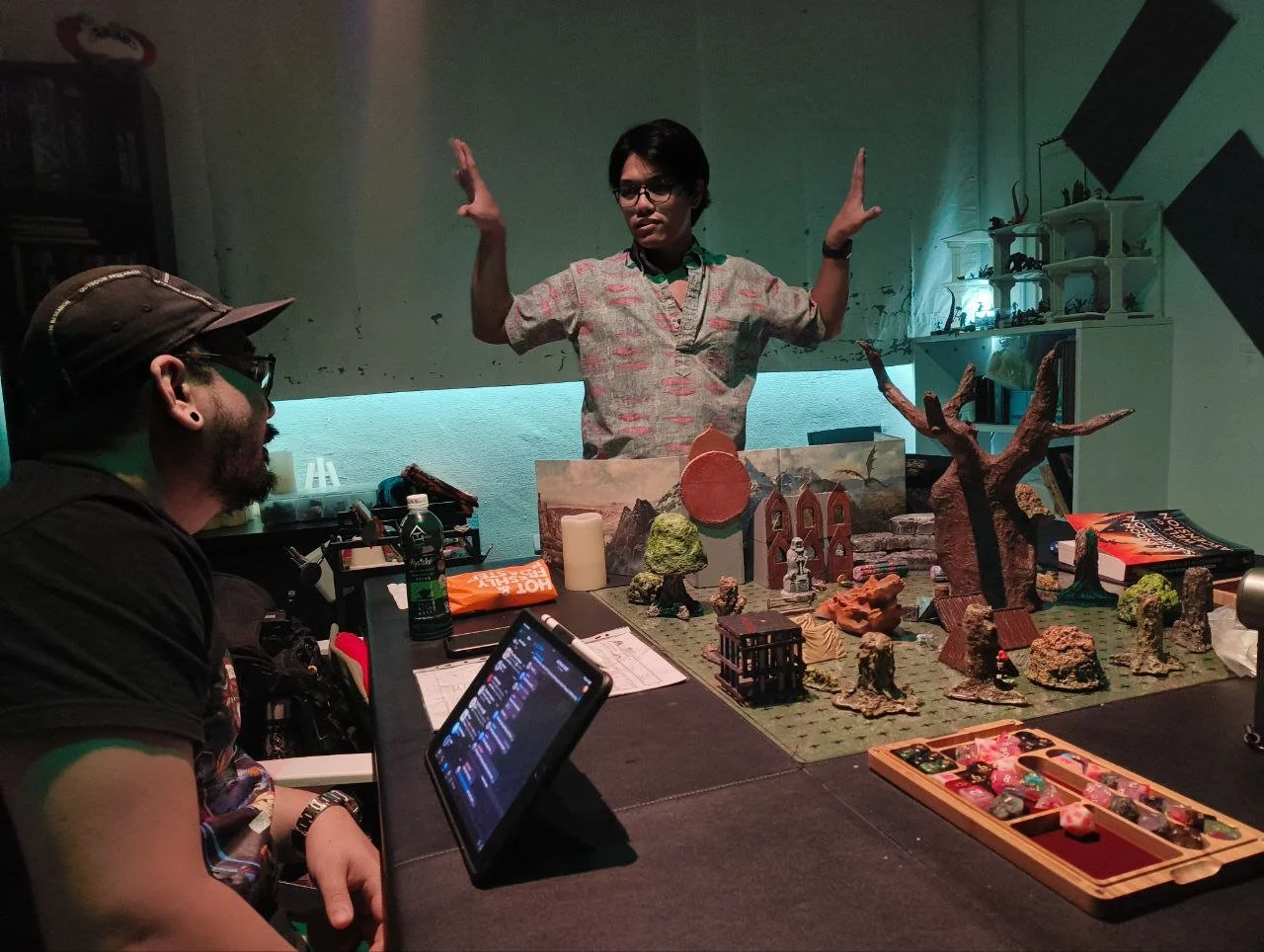 A person with glasses and short dark hair stands behind a tabletop gaming setup with miniature figures and terrain, gesturing animatedly while another person with a cap and glasses sits nearby, observing. The setup includes a large faux tree, painted scenery, and various game components.