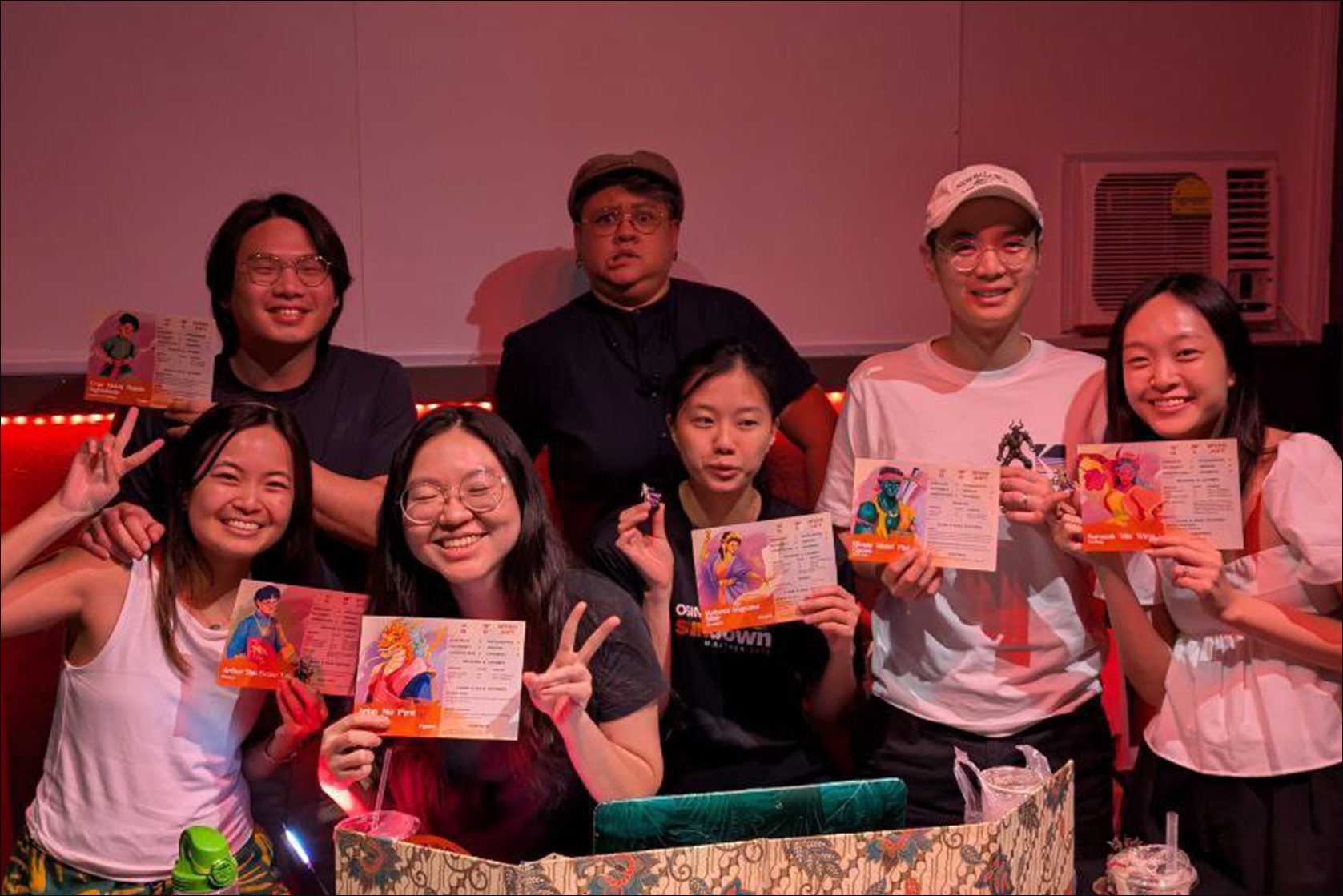 Group of eight young adults holding colorful menus, smiling, and making peace signs inside a restaurant with red lighting.
