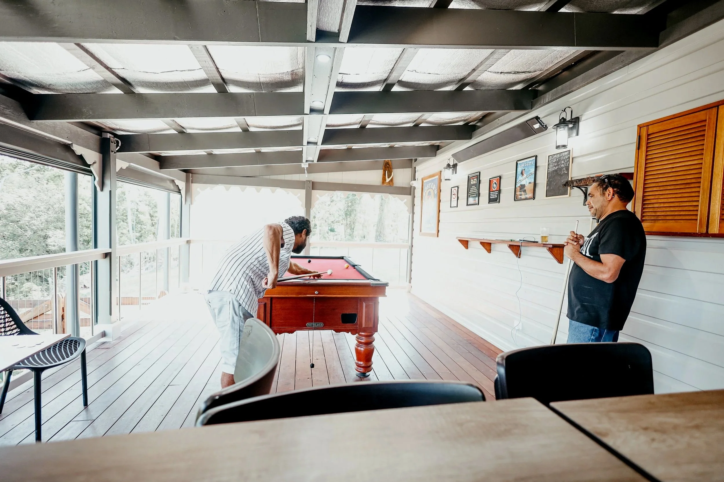 Two men playing pool on a red felt table in a covered outdoor patio area with wooden flooring and a decorative wall with signs and pictures.