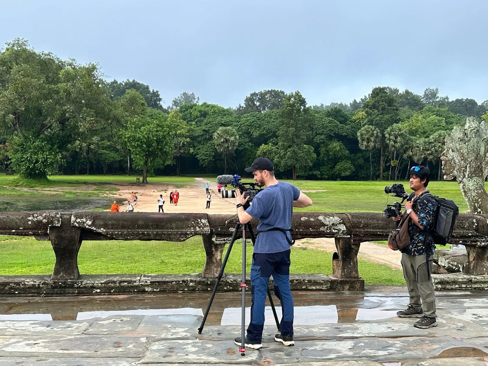 Two videographers filming in front of an old stone bridge with a park and several people in the background.