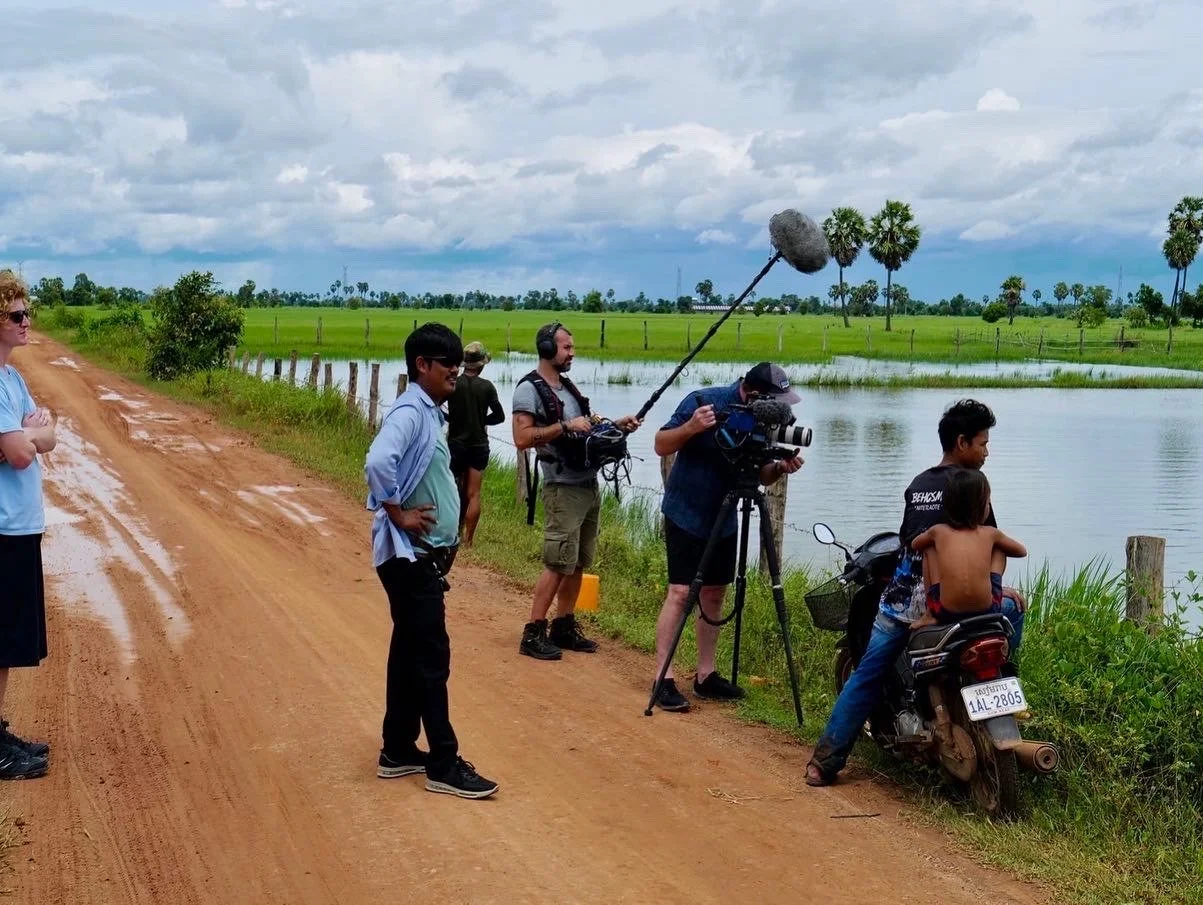 Filming team recording a man on a motorcycle by a river on a dirt road in a rural area with palm trees and green fields under cloudy sky.