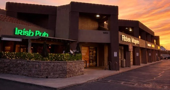 Commercial building with signs for Irish Pub and Fibber Magees, outdoor patio area with flowers and umbrellas, during sunset.