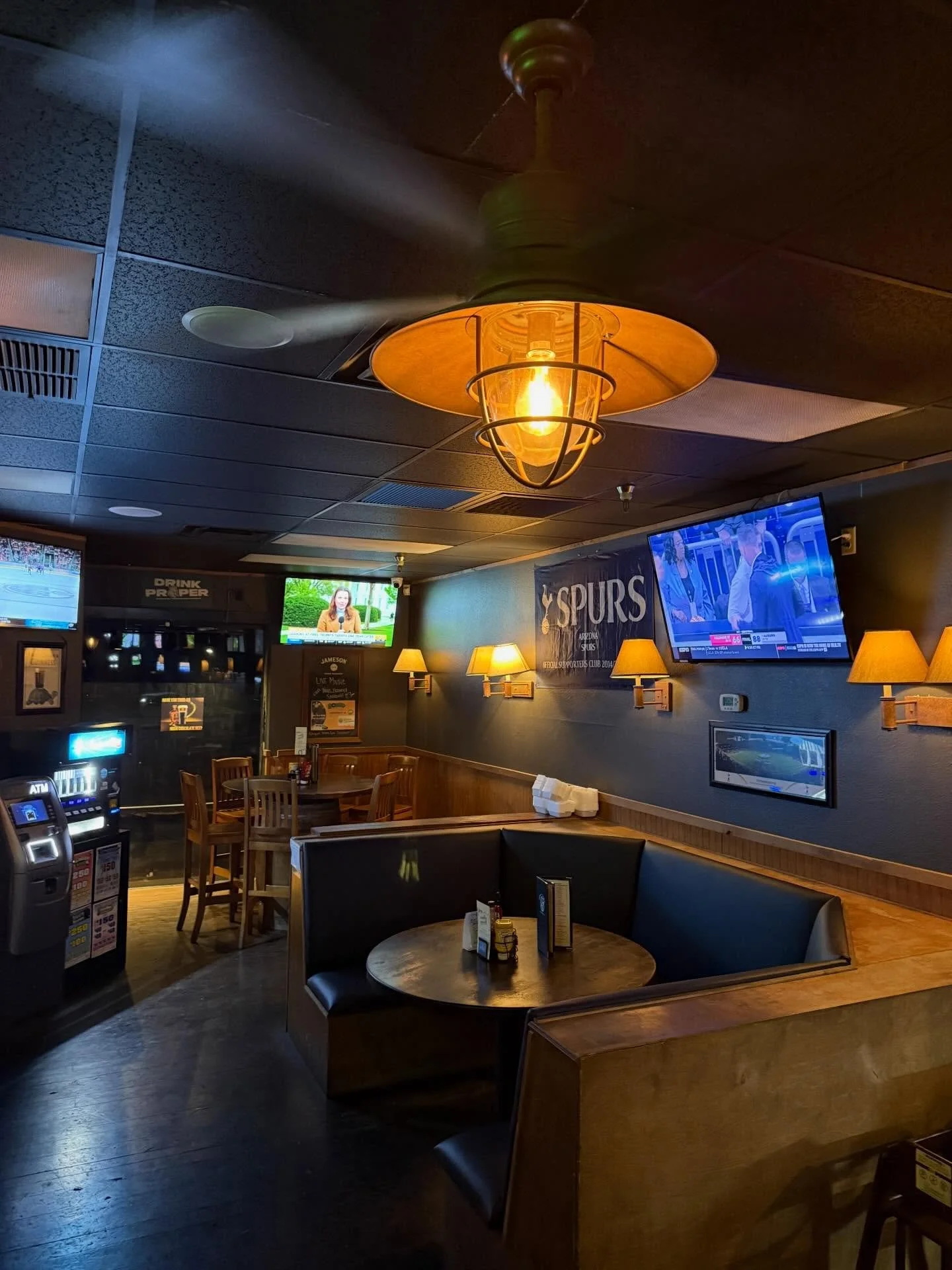 Inside a sports bar with multiple TVs showing a basketball game, wooden tables and chairs, a round booth seat, wall lamps, and a ceiling fan with a glowing light.