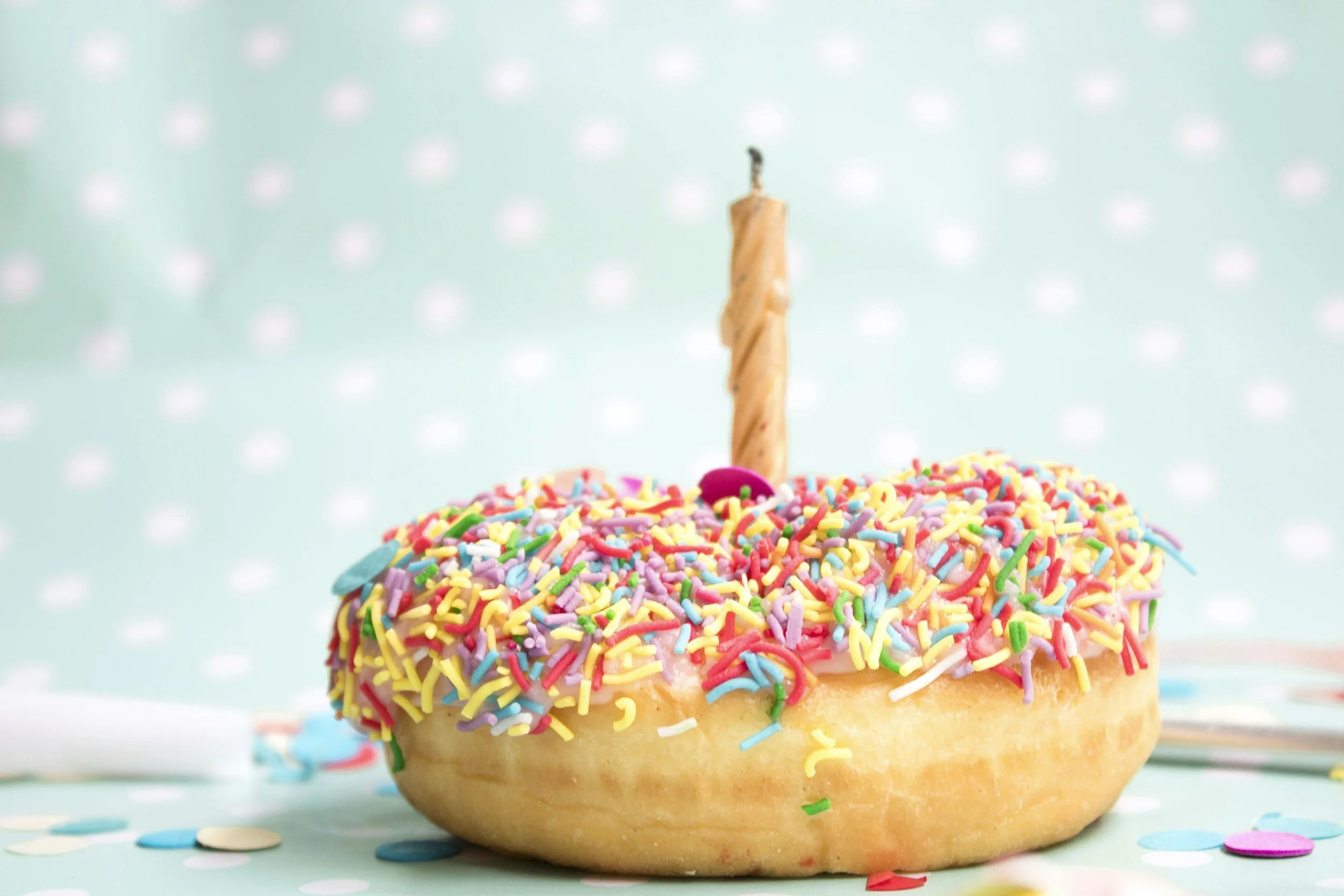 A donut with pink frosting topped with colorful sprinkles and a single lit birthday candle.