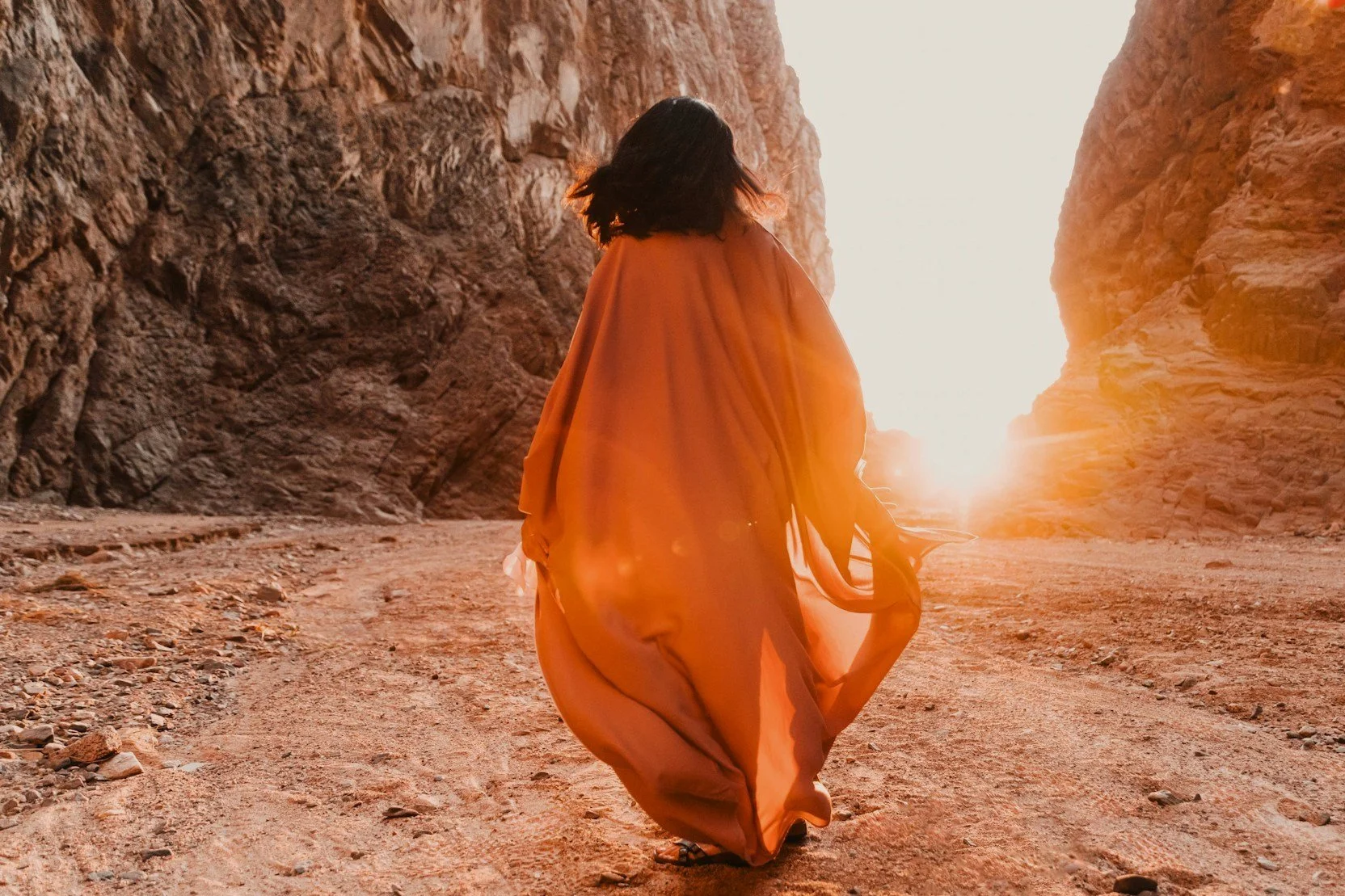 Woman in a flowing orange dress walking through a sunlit rocky canyon