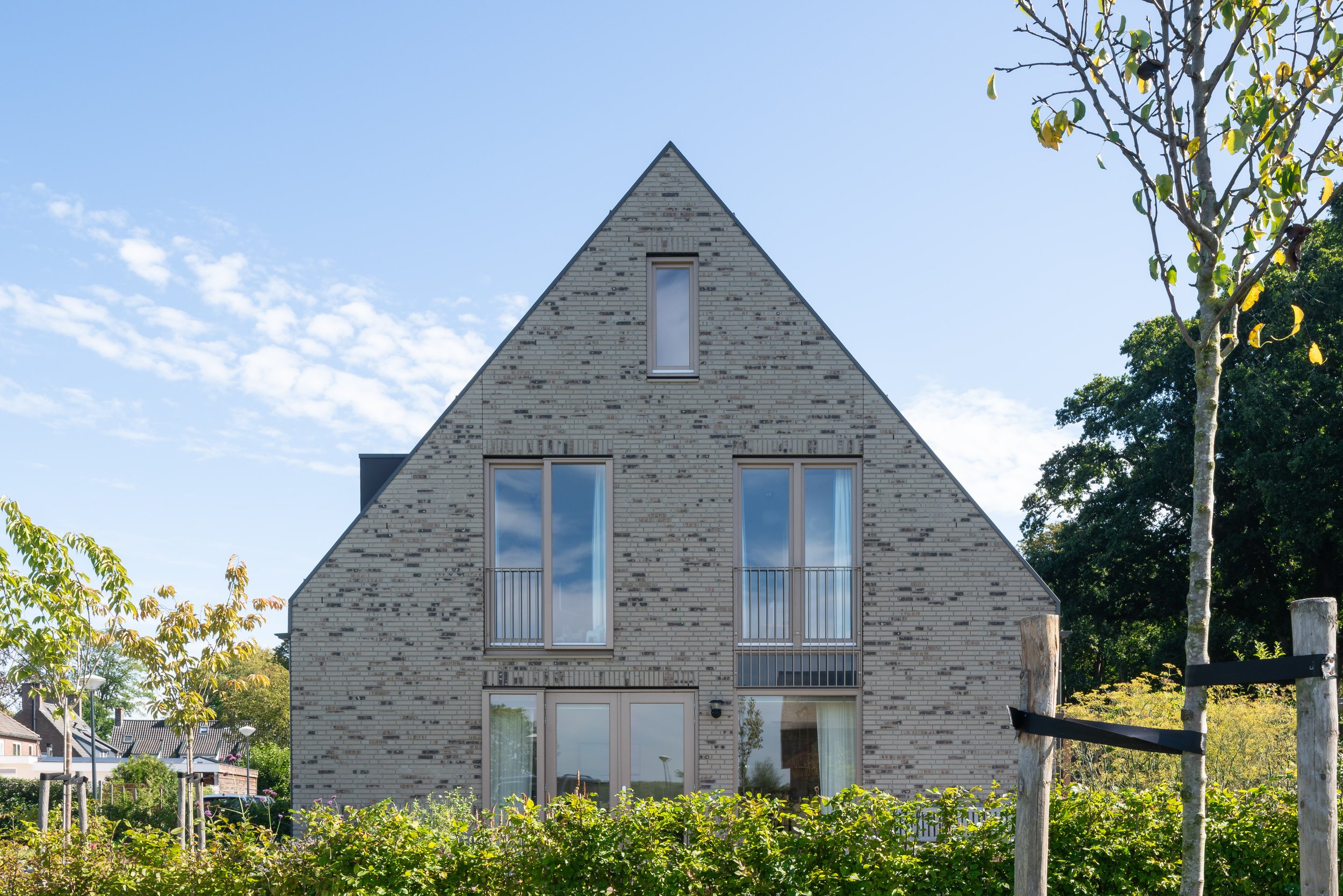 Straataanzicht van een nieuwbouwvilla met een lichte gemêleerde baksteengevel en een spitse topgevel met dakraam en balkon in Nederhorst den Berg als woningbouw architectuur.