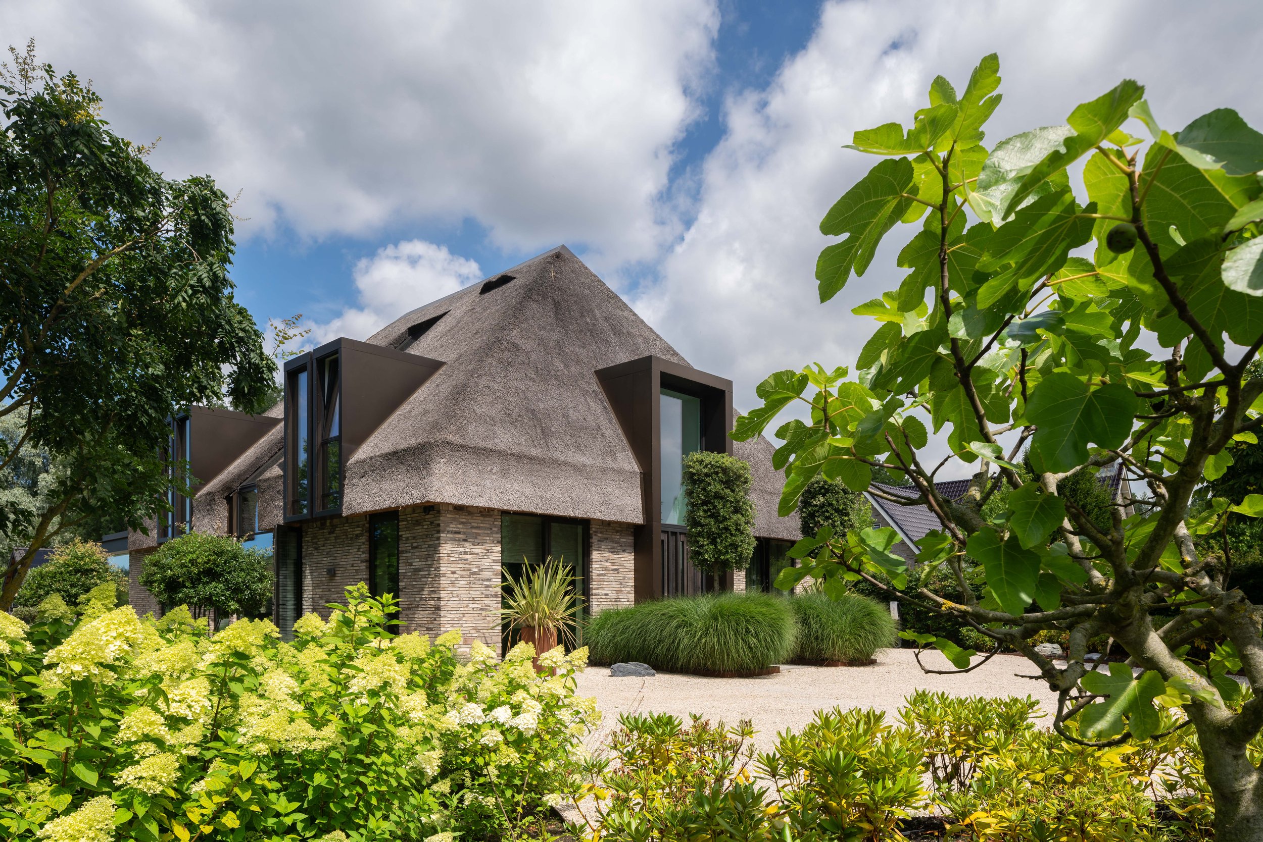 Moderne woning met rieten dak, omgeven door groene planten en bomen onder een bewolkte blauwe hemel.