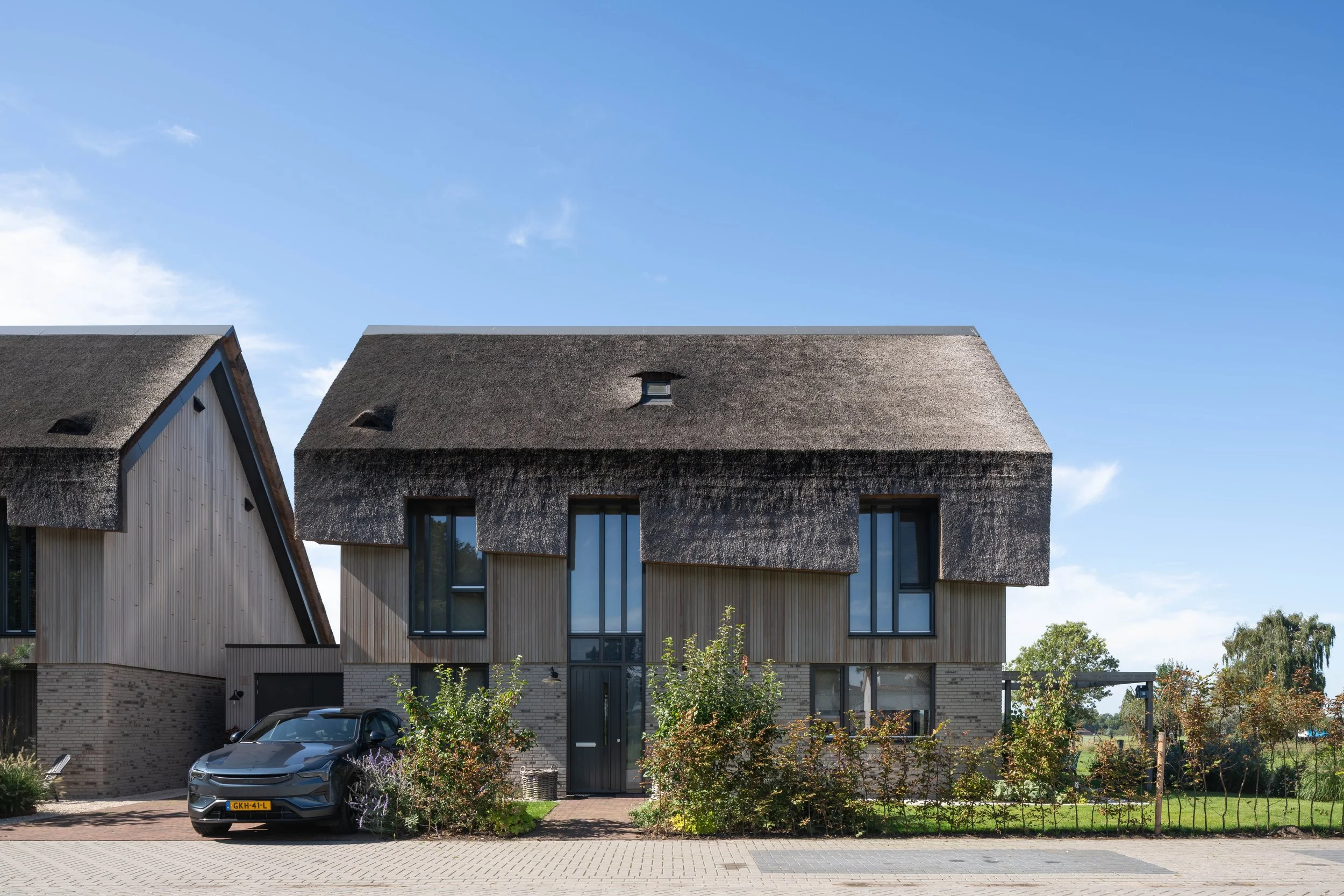 Vooraanzicht van een vrijstaande nieuwbouwwoning met een rietgedekt dak, houten gevelbekleding en een lichte bakstenen plint met grote glaspartijen in Nederhorst den Berg.