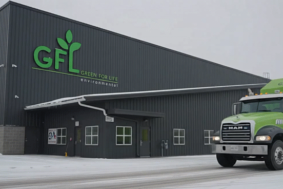 A bright green GFL Environmental truck is parked outside a dark industrial building with the company's large green logo mounted on the wall. Snow covers the ground around the vehicle.