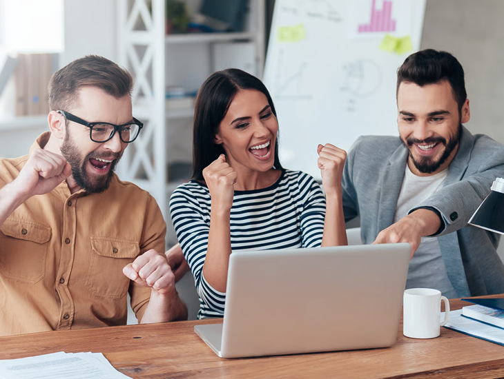Three smiling office colleagues celebrate a win around a laptop.