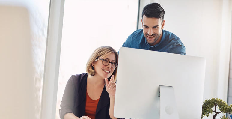 Two employees, one seated and one standing, viewing a desktop monitor.