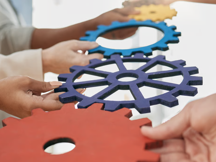 A close-up of multiple hands holding and connecting large, colorful cardboard gears together.