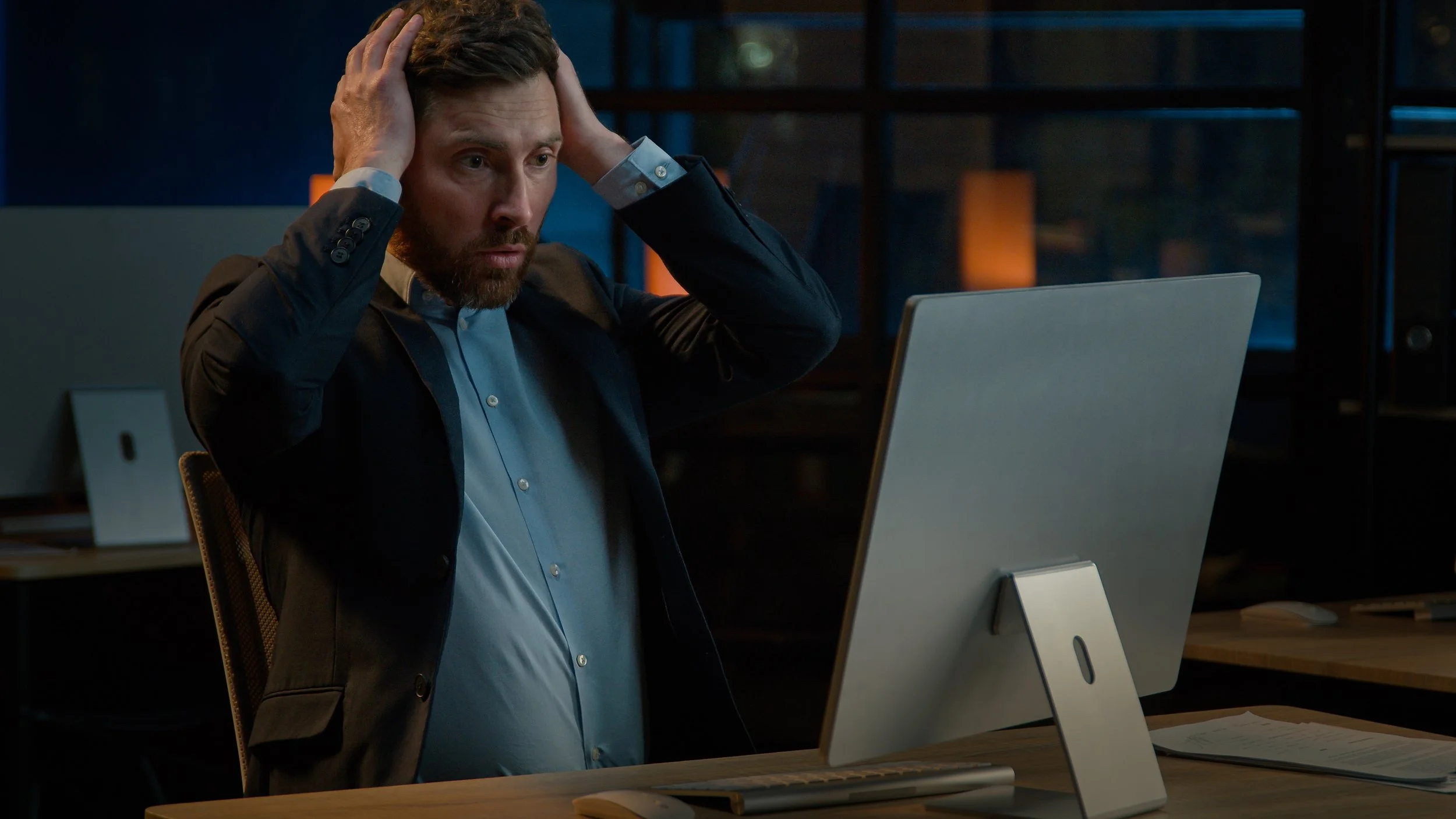 A businessman working late at night in a dark office, staring at his computer screen with a shocked and frustrated expression, holding both hands to his head.