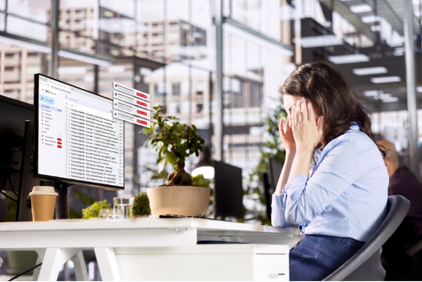 A frustrated woman sitting at a desk in an office, holding her head in her hands in front of a computer monitor displaying an email inbox.