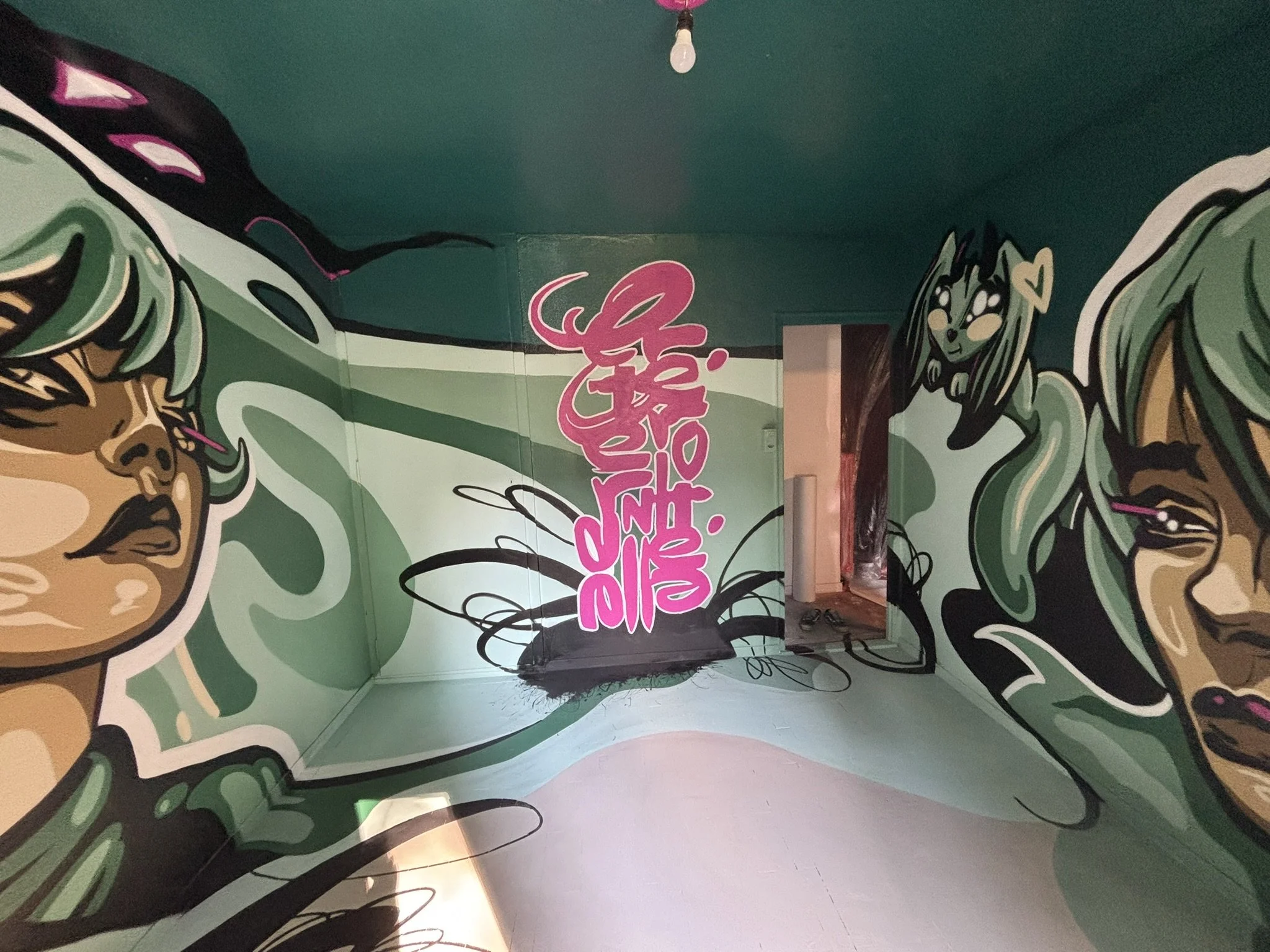 Interior of a room with graffiti-style mural art on the walls, featuring stylized portraits of two women with green hair, and bold pink graffiti text in the center.