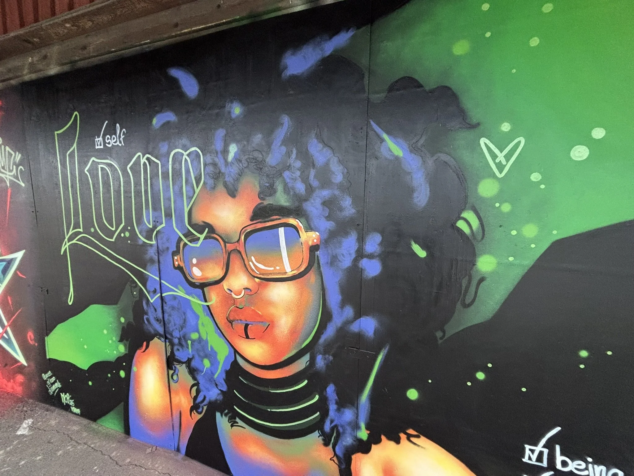 Colorful graffiti mural of a woman with curly hair, wearing large sunglasses, a nose ring, and a choker, with vibrant green, blue, and black backgrounds and spray paint effects.
