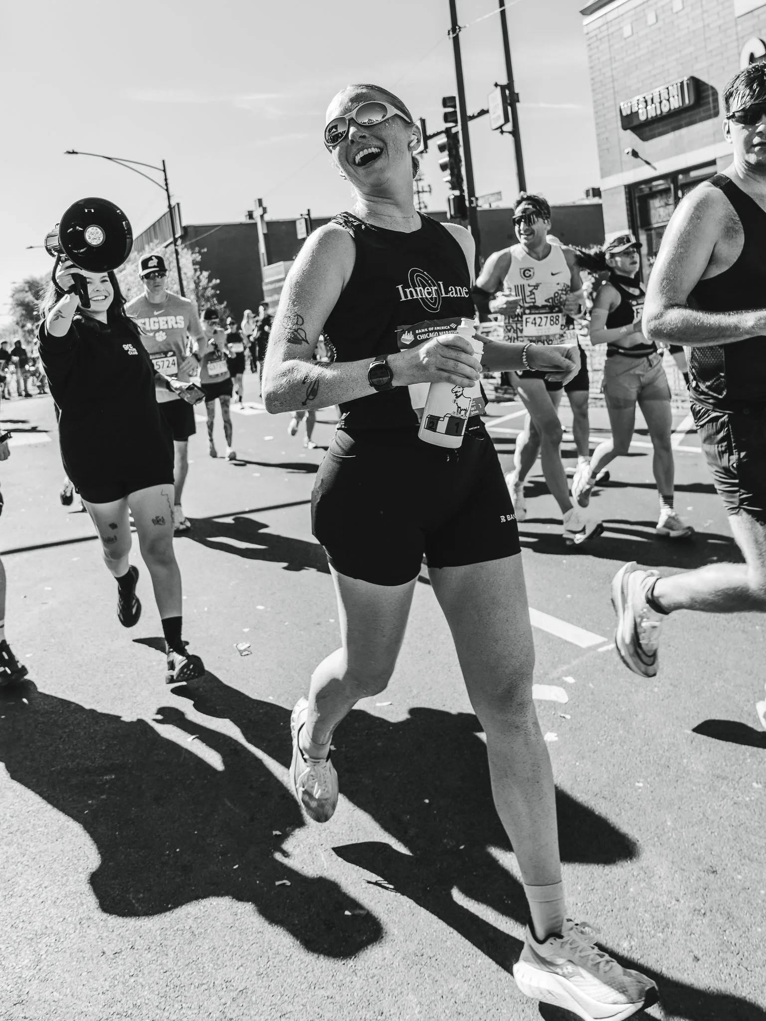Black and white photo of marathon runners on city street, including a woman with sunglasses and a tank top, some smiling and talking, with streetlights and building in the background.
