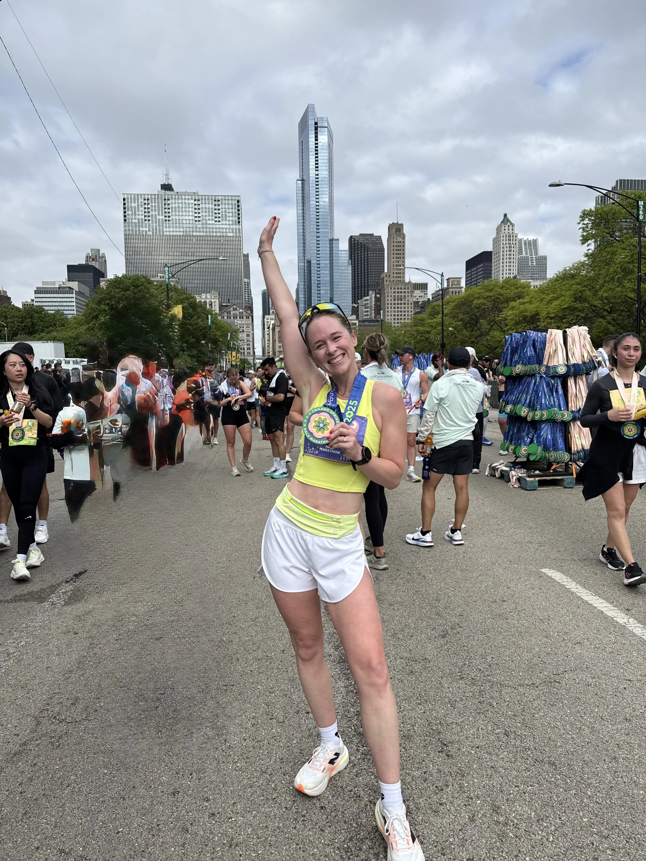 A smiling woman posing with one arm raised in front of a city skyline with tall buildings, wearing running gear, a medal around her neck, and surrounded by other runners and street vendors.