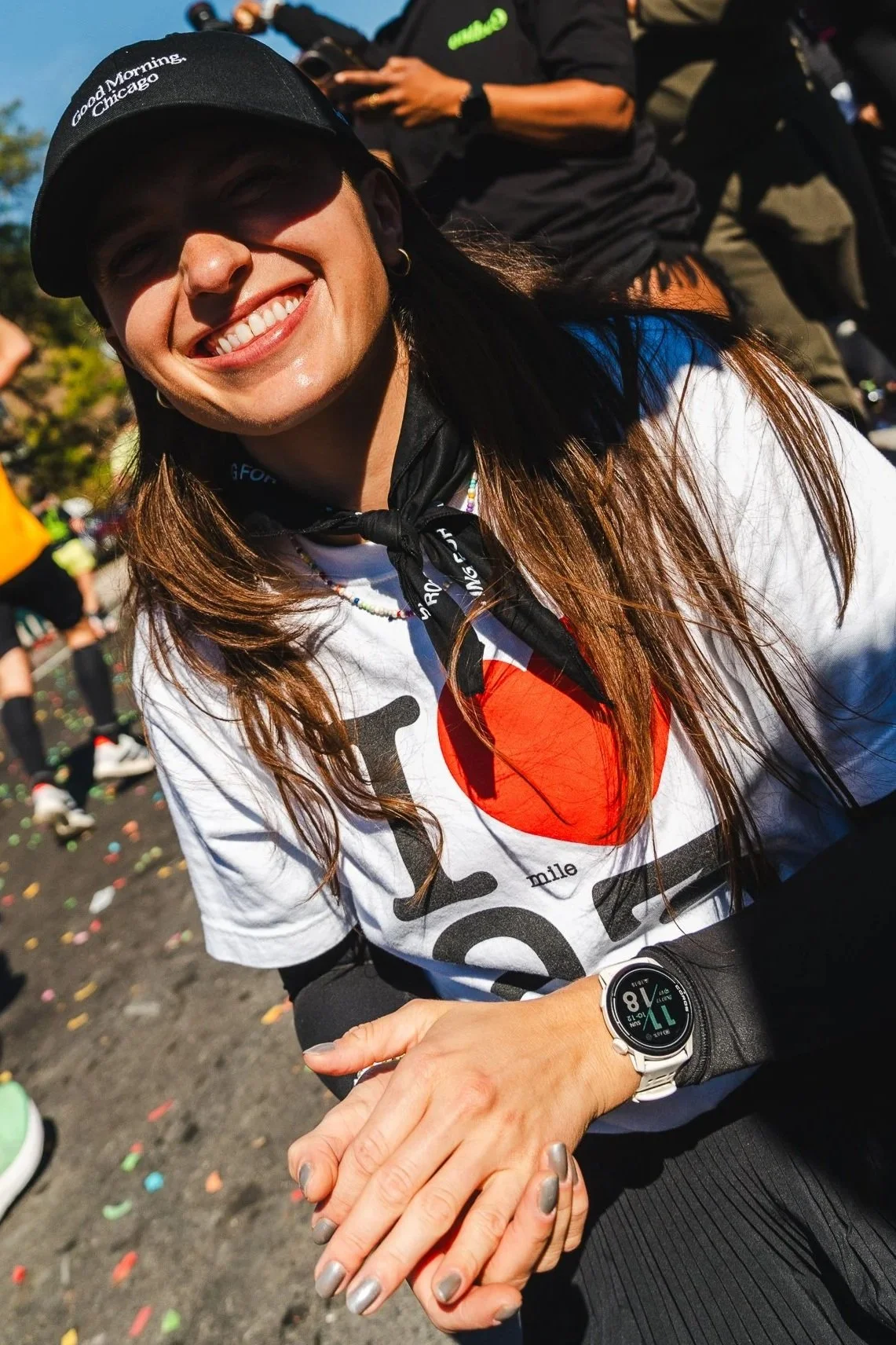 Smiling woman outdoors wearing a black cap, white t-shirt, and a black jacket tied around her neck. She is holding hands with another person. Several people and colorful confetti are in the background.