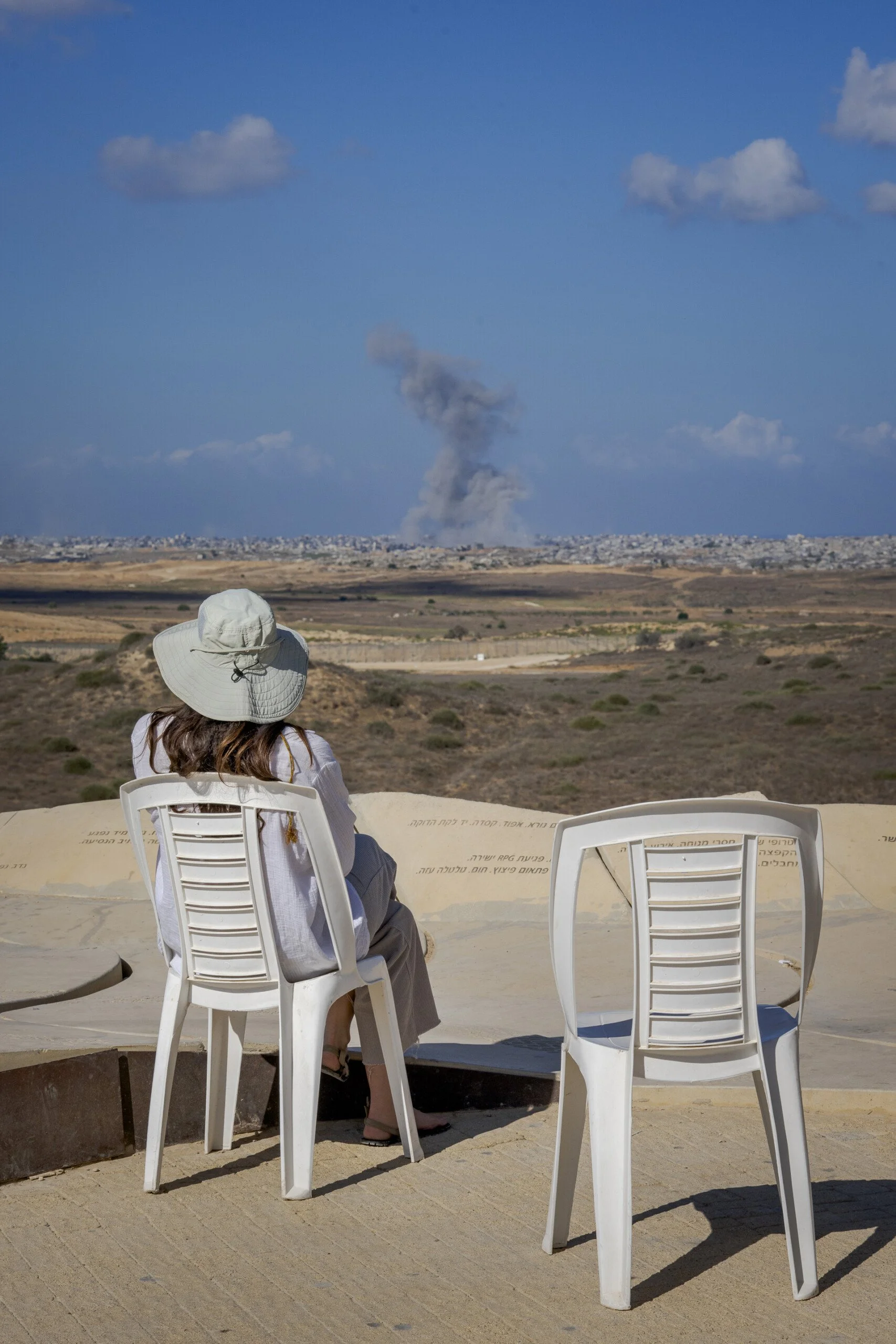 En kvinna i solhatt sitter i en vit plaststol med ryggen mot kameran, blickandes mot en rökpelare i horisonten. Tre kilometer framför henne ligger norra Gaza.