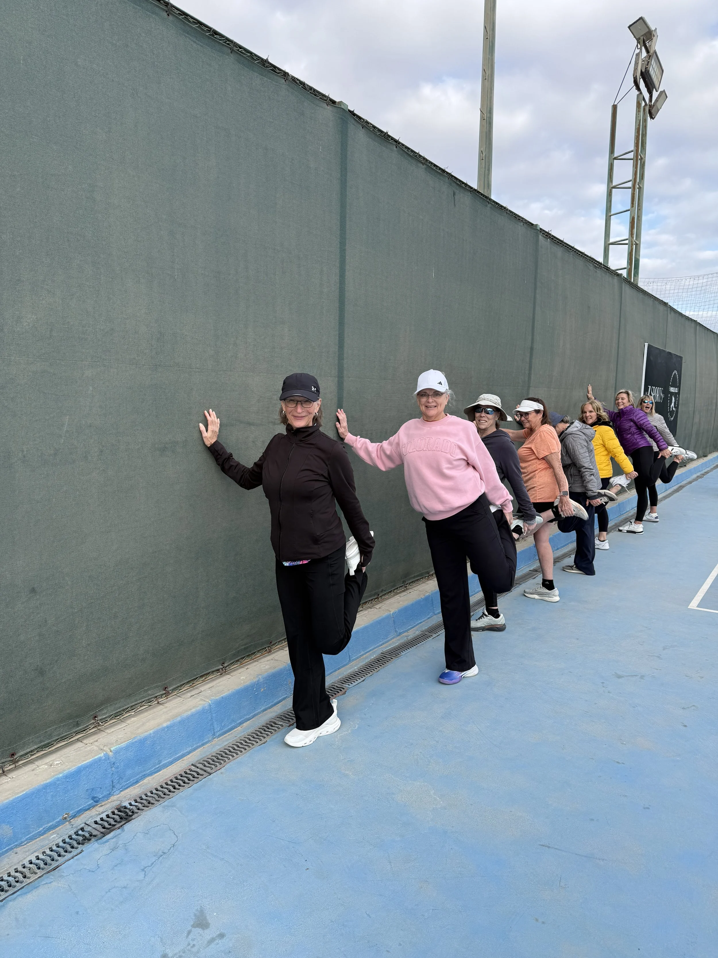 Group of women practicing yoga outdoors, performing a stretch with one leg bent and their hands on the wall, on a blue tennis court surface.