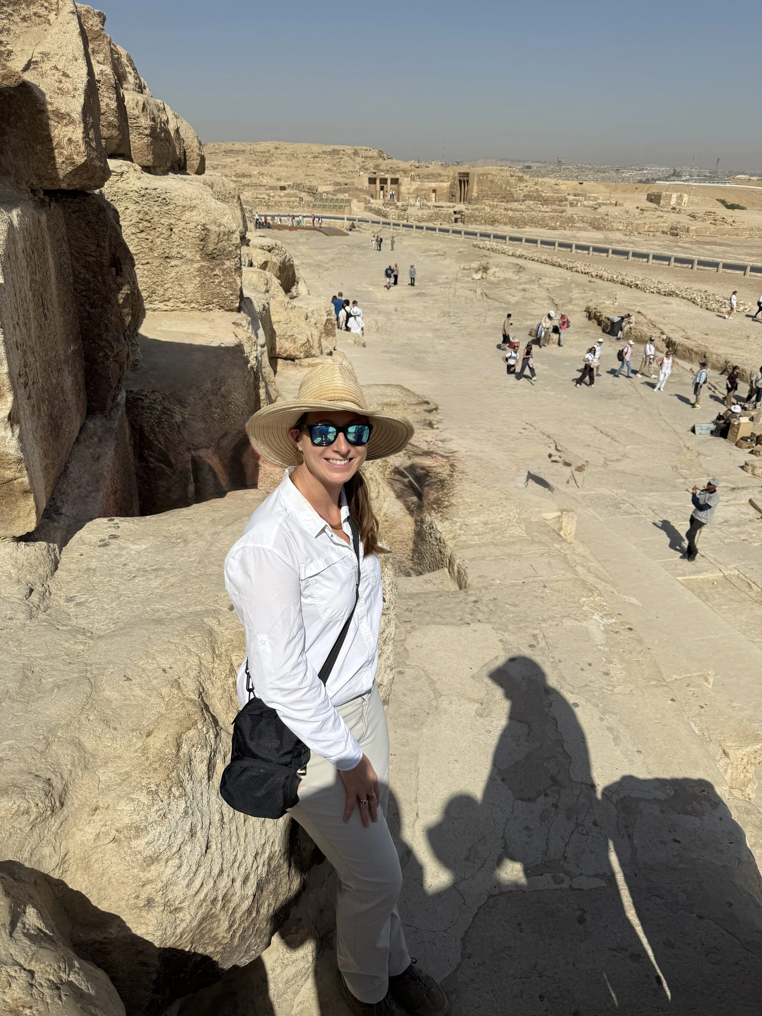 A woman in a white blouse and straw hat wearing sunglasses stands on a side of the Great Pyramid of Giza.