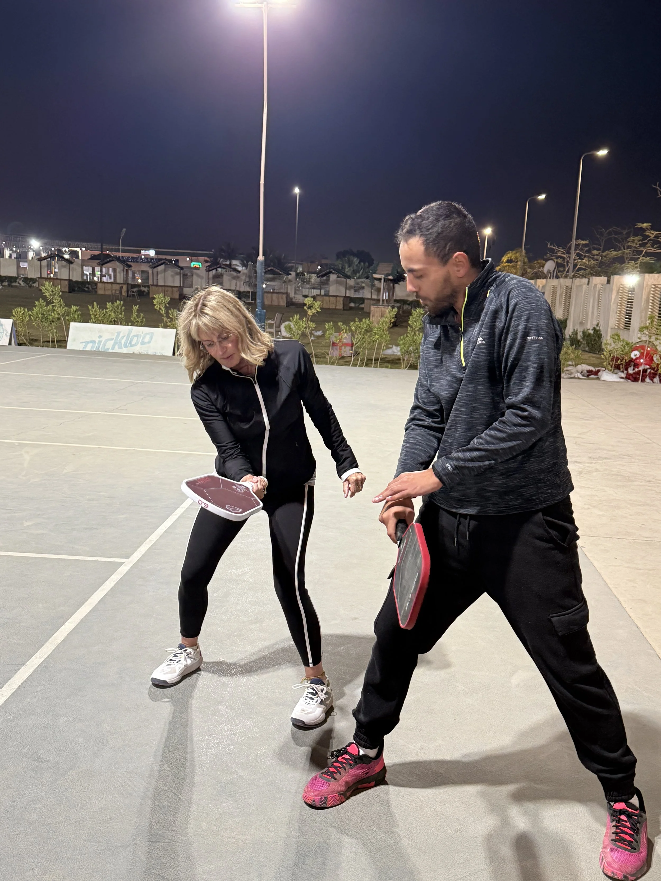Two people playing pickleball on an outdoor court at night, with a woman preparing to hit the ball and a man holding a paddle, illuminated by overhead lights.