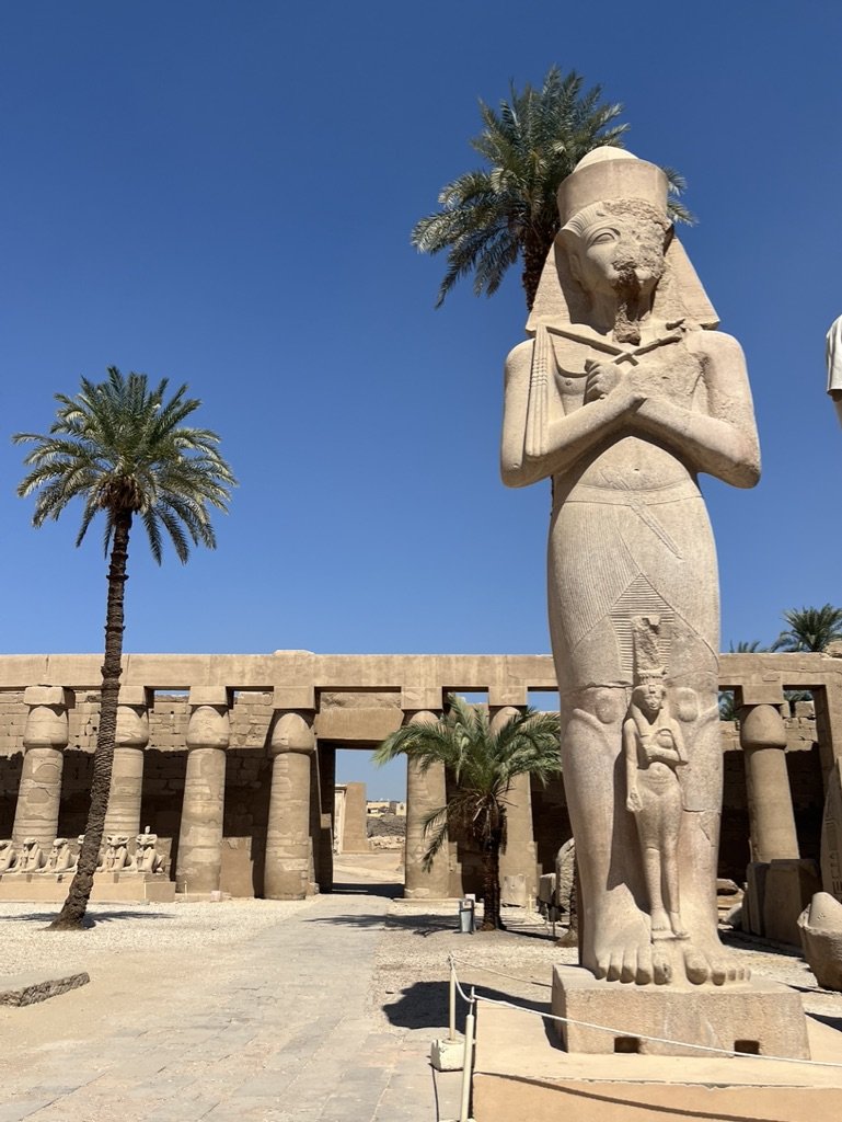 Ancient Egyptian statue in Karnak Temple complex, Luxor, with palm trees and ruins in the background under a clear blue sky.