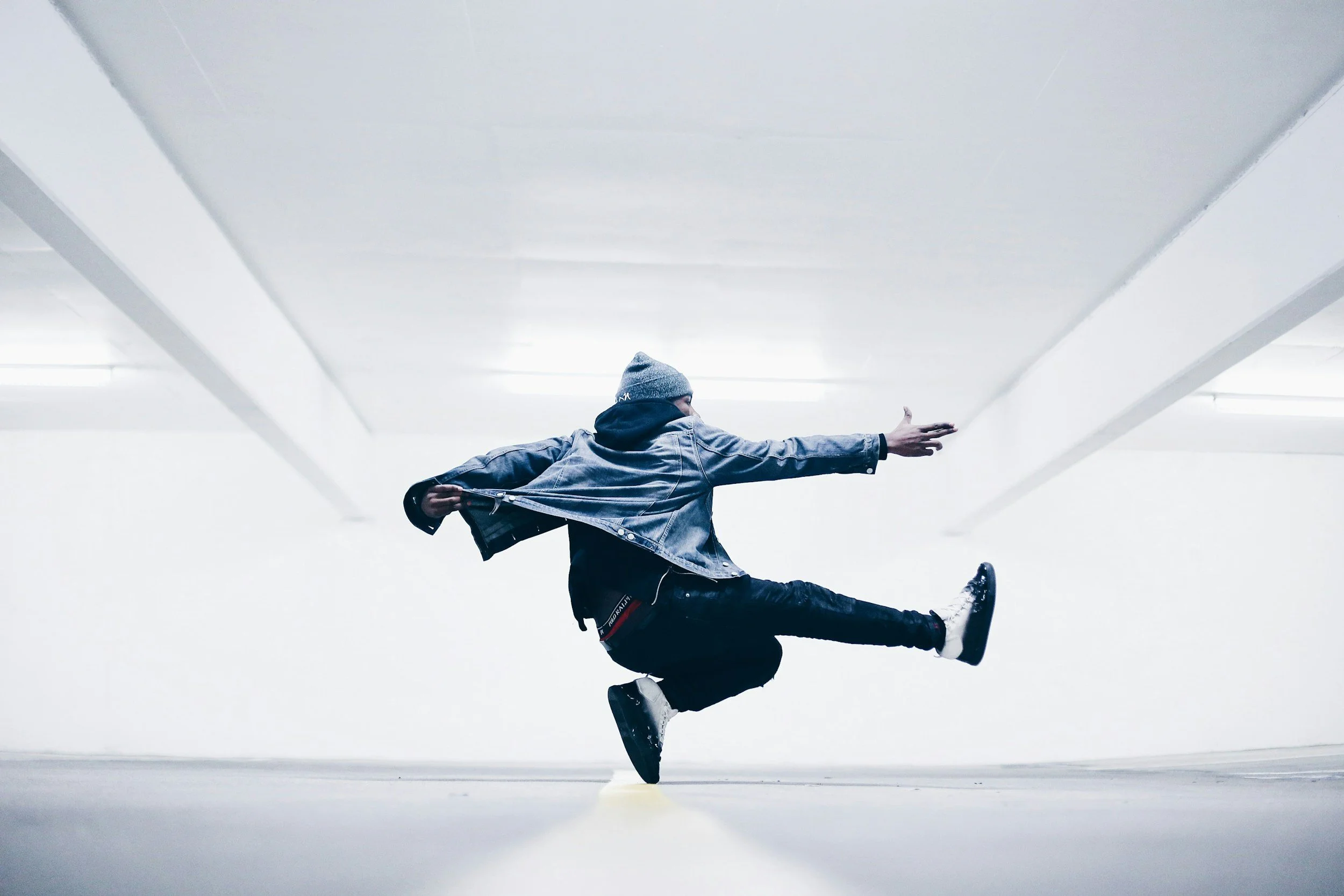 A person dressed in a grey hoodie, denim jacket, and black pants jumps in the air, striking a pose with one arm extended forward and one leg extended backward in a minimalist, empty parking garage.