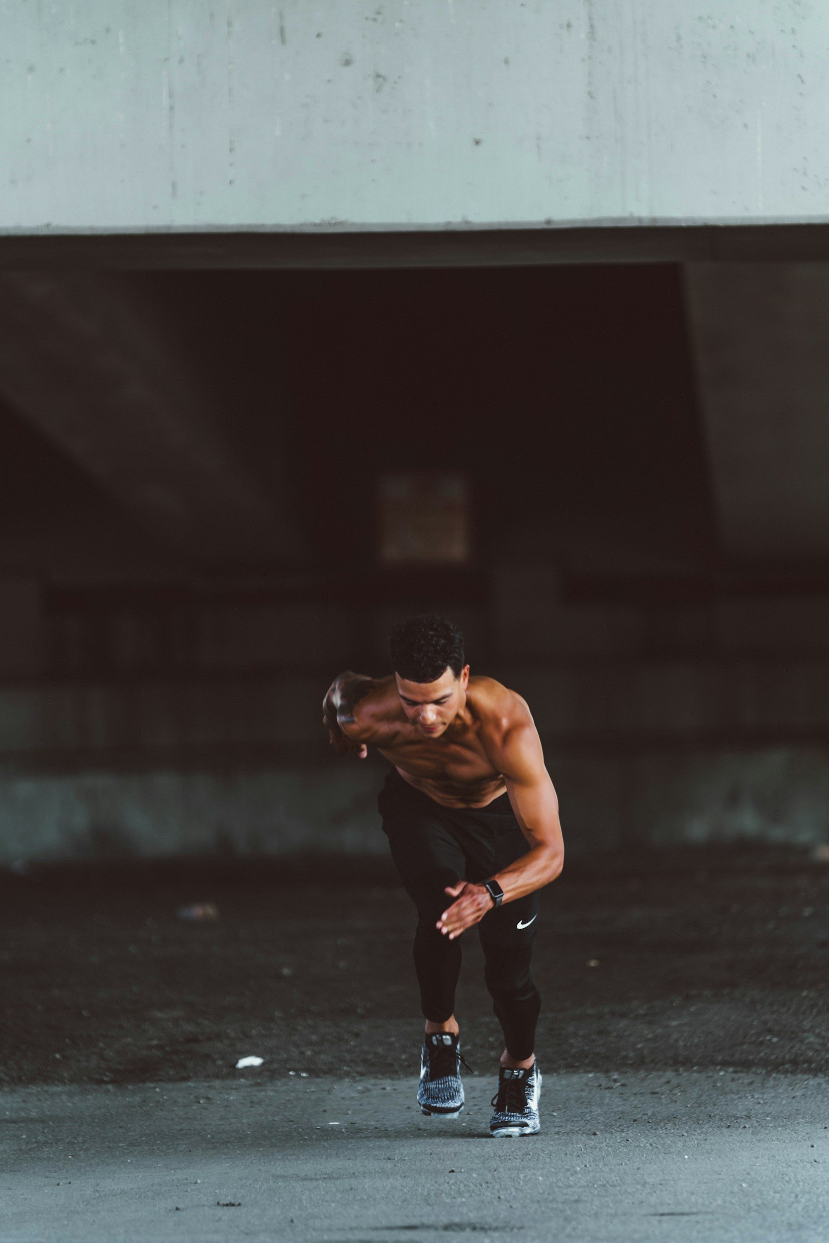 A shirtless man running under a bridge, wearing black pants and running shoes.
