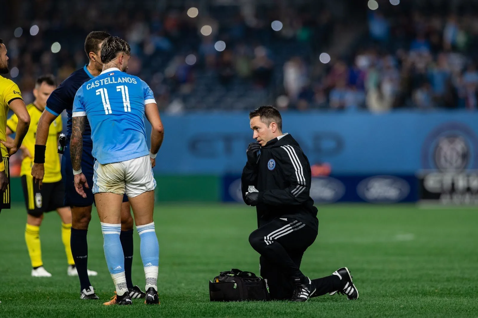 A soccer coach kneeling on the field speaking to a player in a blue jersey, with other players and a crowd in the background.