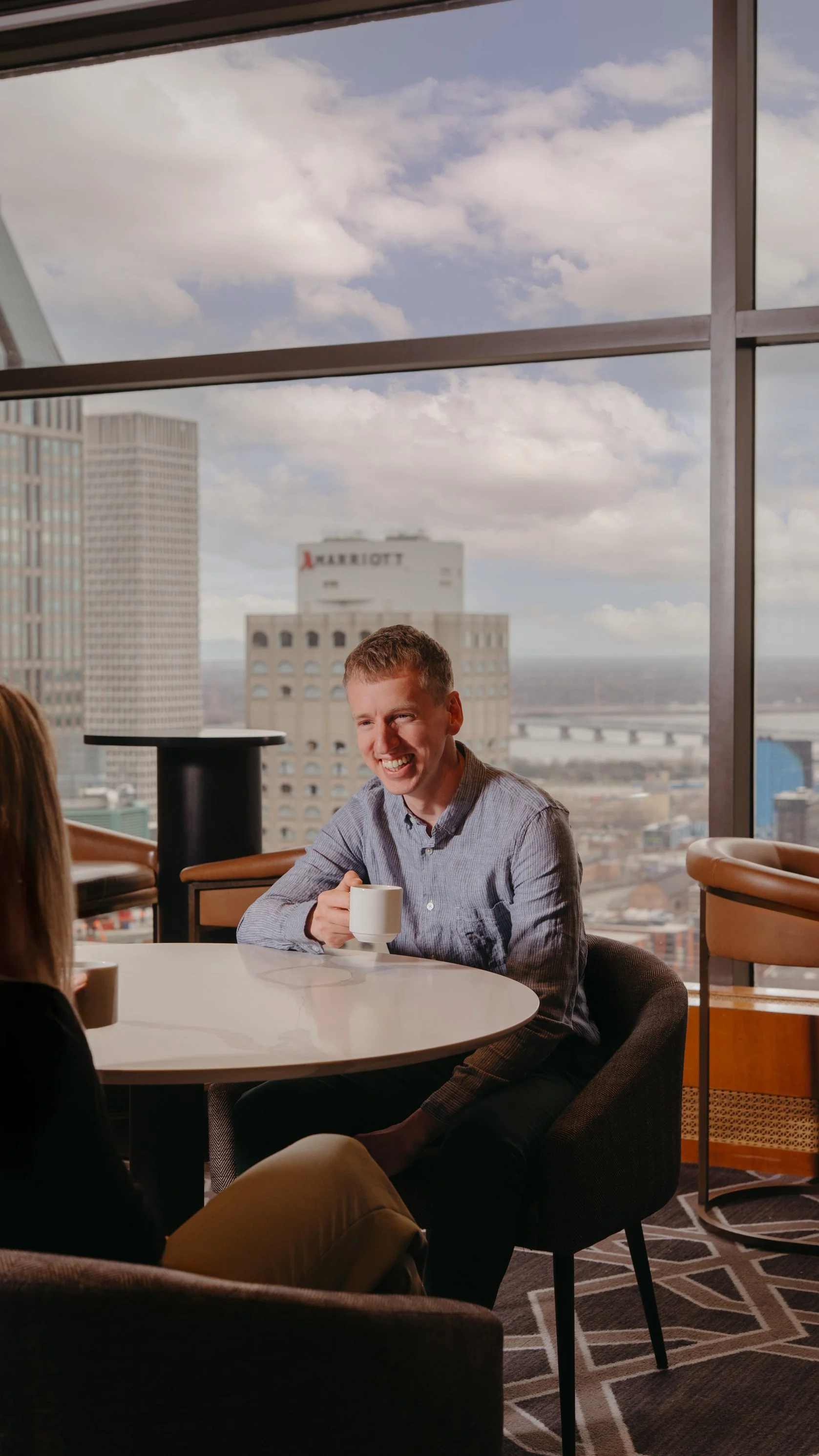 A man smiling and sitting at a table with a cup in hand in a modern office with large window showing city buildings and partly cloudy sky.