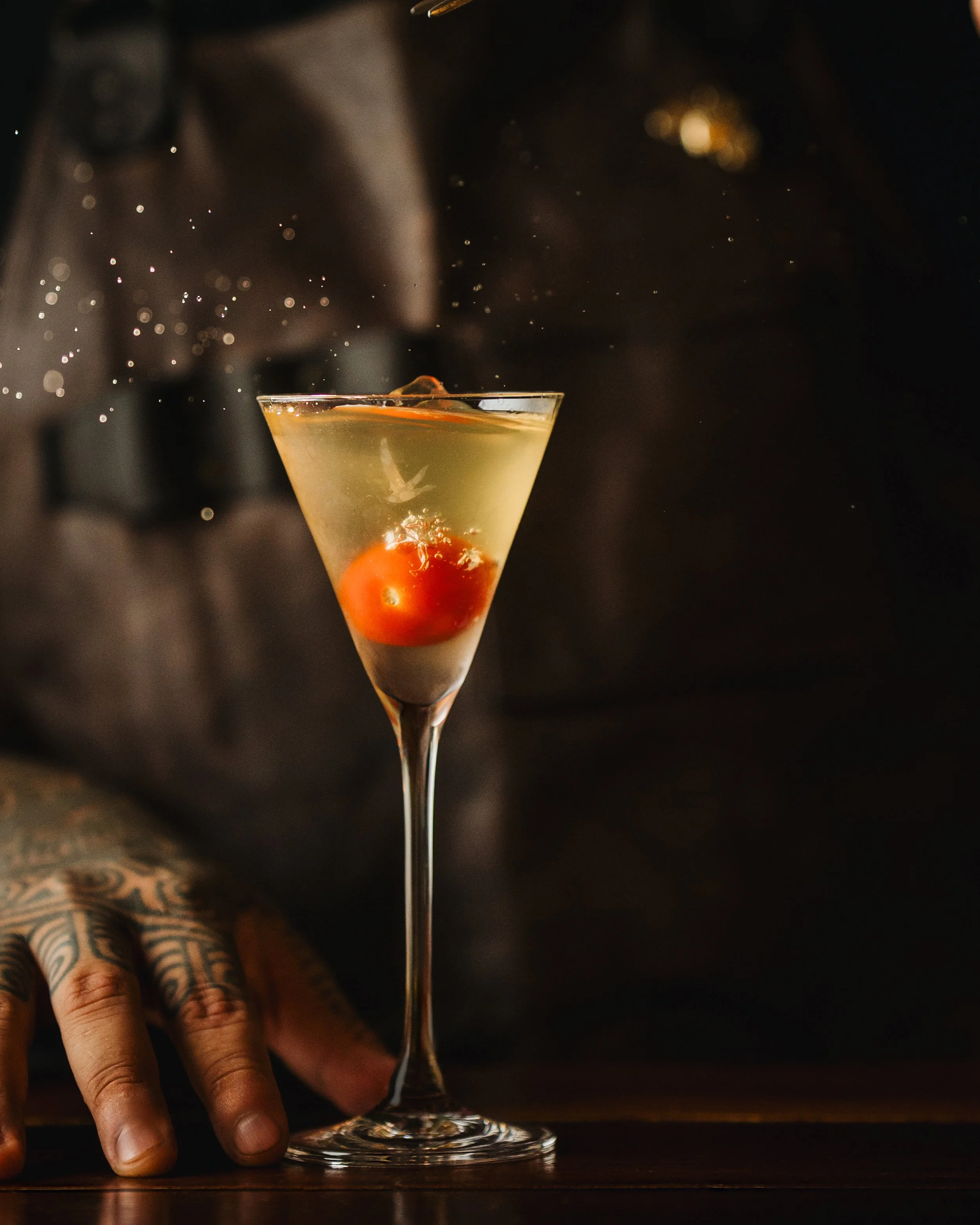 A close-up of a person's hand with tattoos resting on a wooden surface, with a martini glass filled with a yellowish drink and garnished with a cherry and an orange slice, against a dark background.