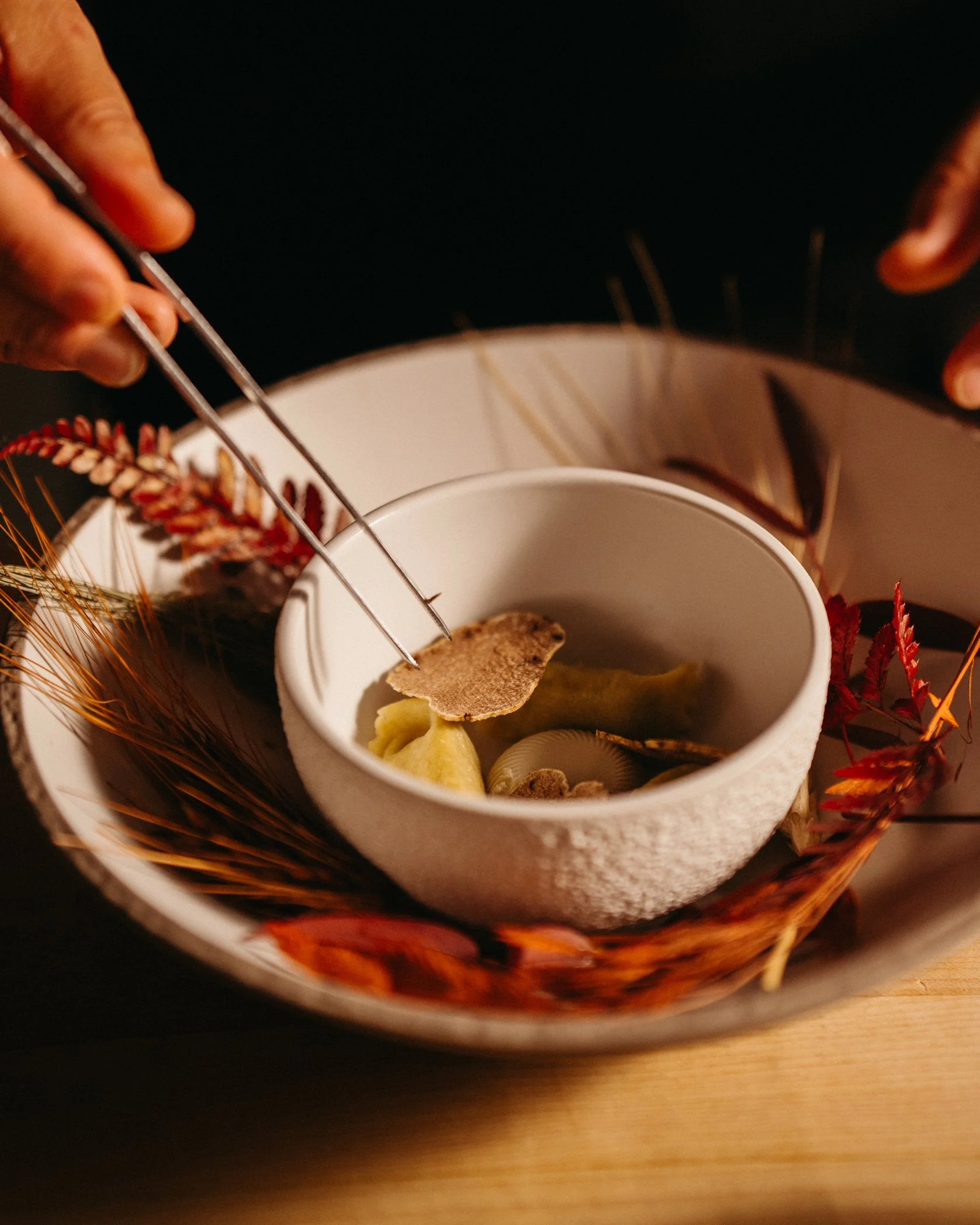 Close-up of a white mug with a hot beverage, garnished with a slice of ginger, inside a woven basket with autumn leaves and decorative dried plants. A person's hands are seen using tongs to pick a piece of ginger from the beverage.