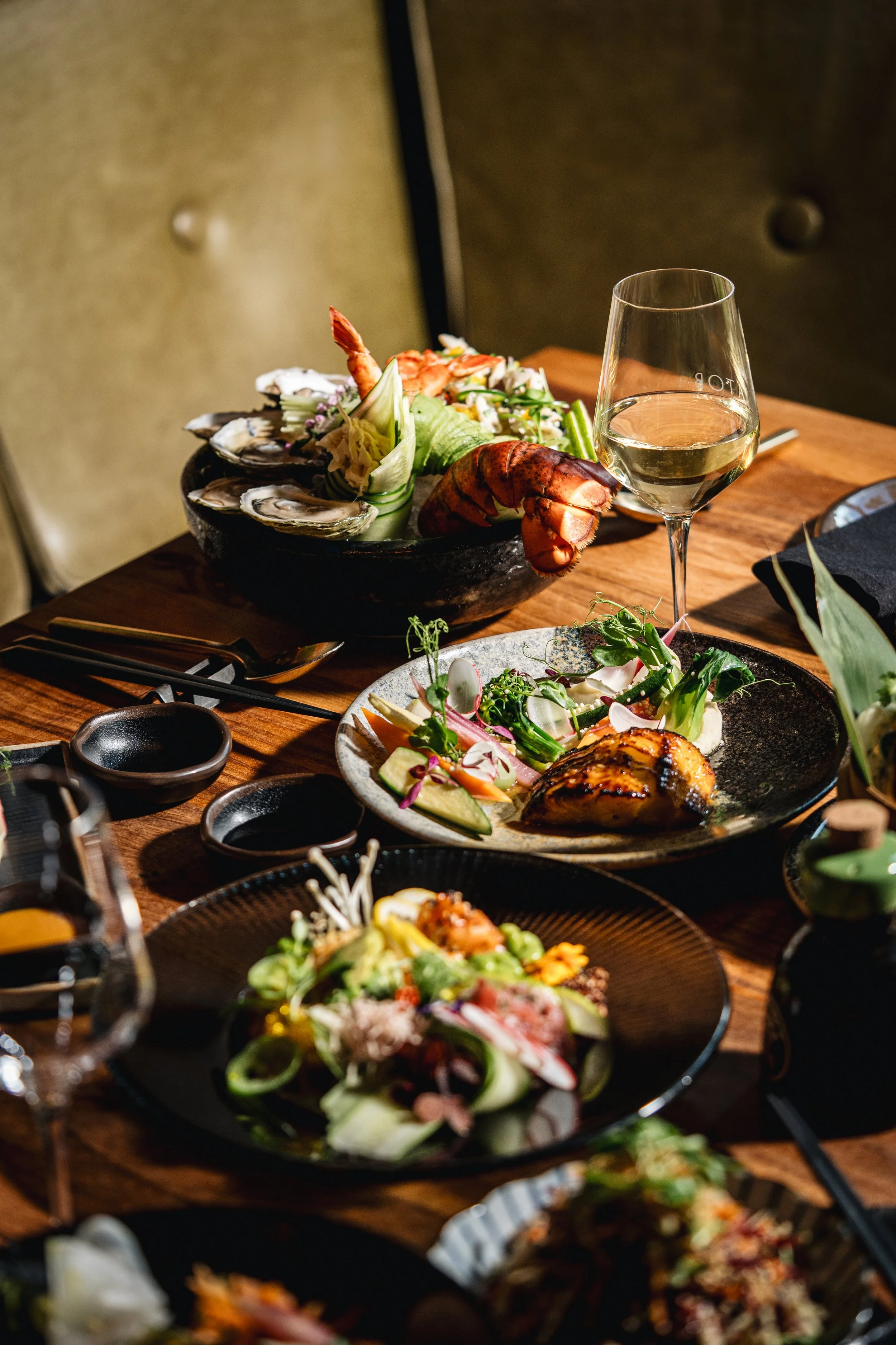 A table set with various dishes including oysters, lobster, salad, and a glass of white wine. The dishes are placed on a wooden table with black plates and utensils.