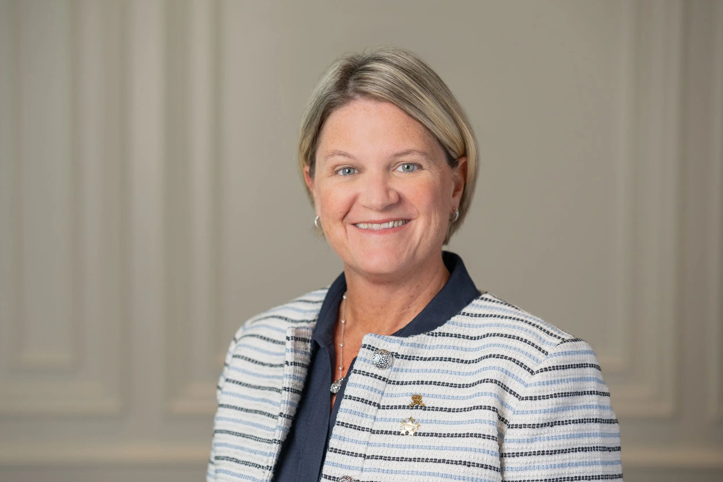 A middle-aged woman with blonde hair, blue eyes, and earrings, smiling at the camera. She is wearing a striped blazer with decorative pins and a dark blouse underneath, standing against a neutral background.