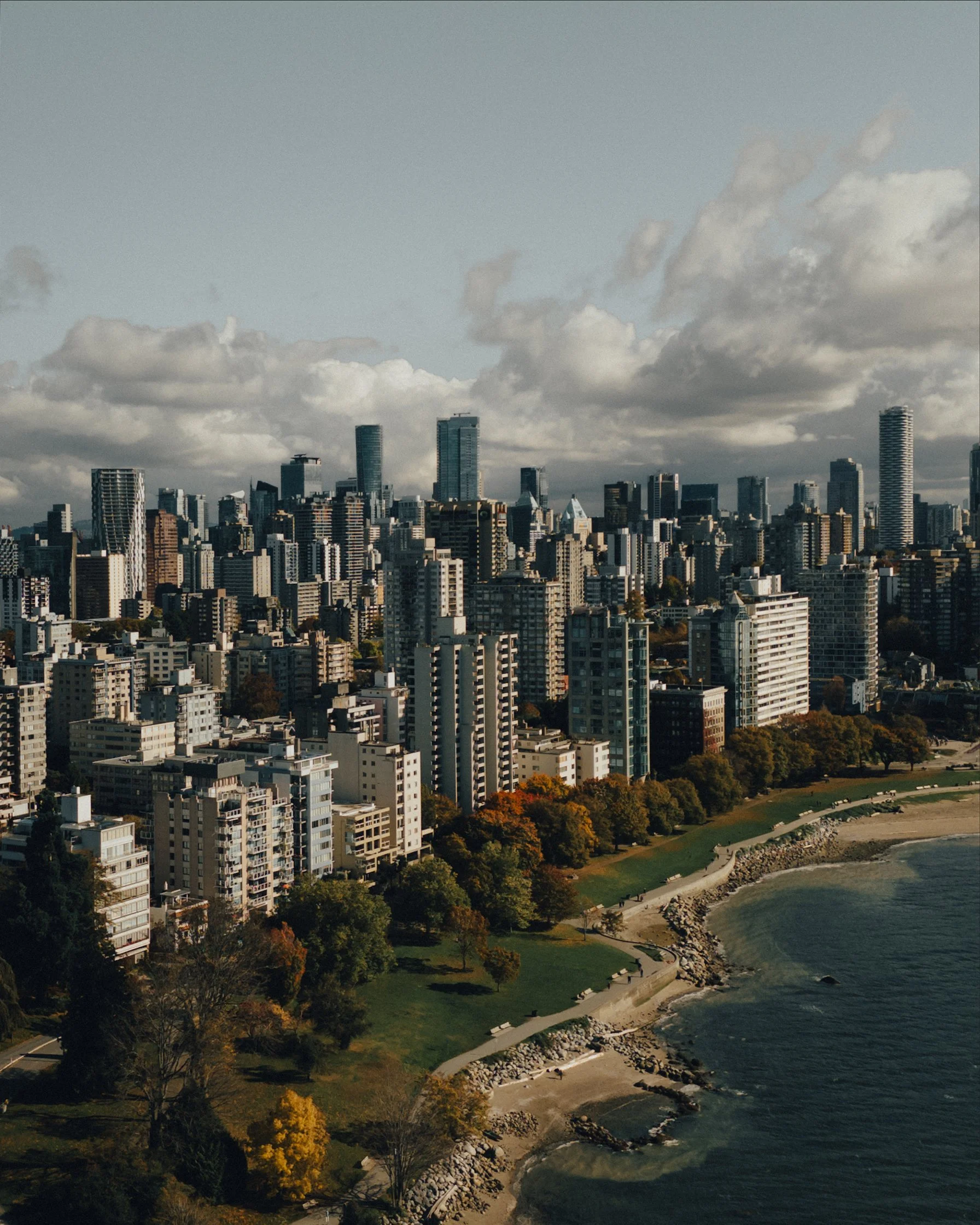 A panoramic view of a city skyline along a coastline with a park and trees in the foreground, under a cloudy sky.