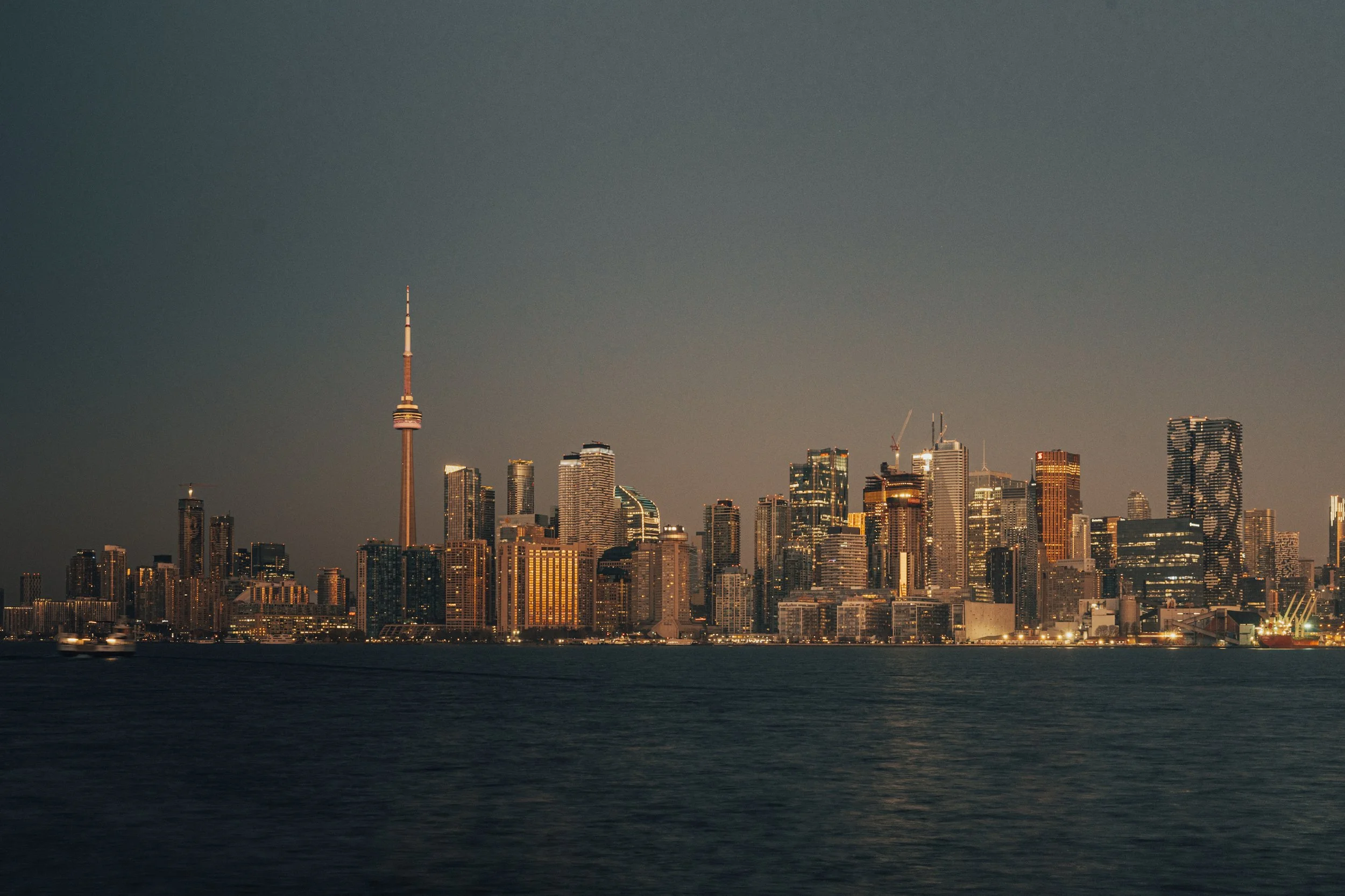 Nighttime city skyline featuring tall buildings and a prominent TV tower reflected in a river or bay.