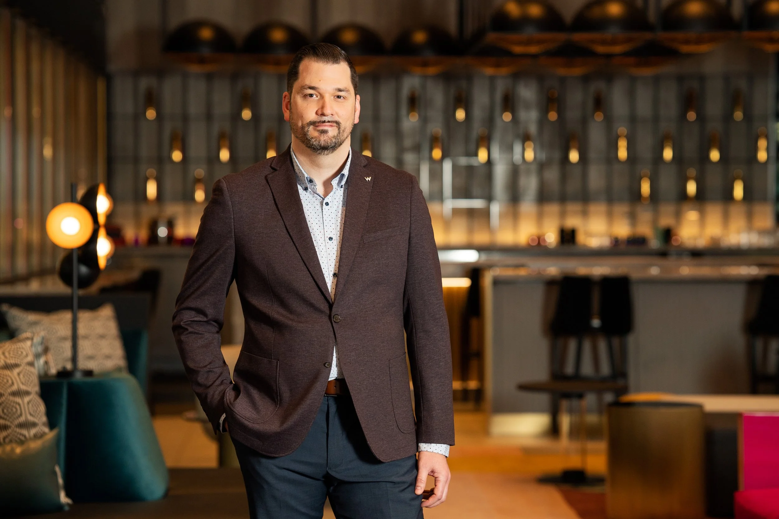 A man with dark hair, a beard, wearing a maroon blazer, a white shirt with small dark dots, and dark pants, standing in a modern interior space with warm lighting, shelves in the background, and seating area with pillows.