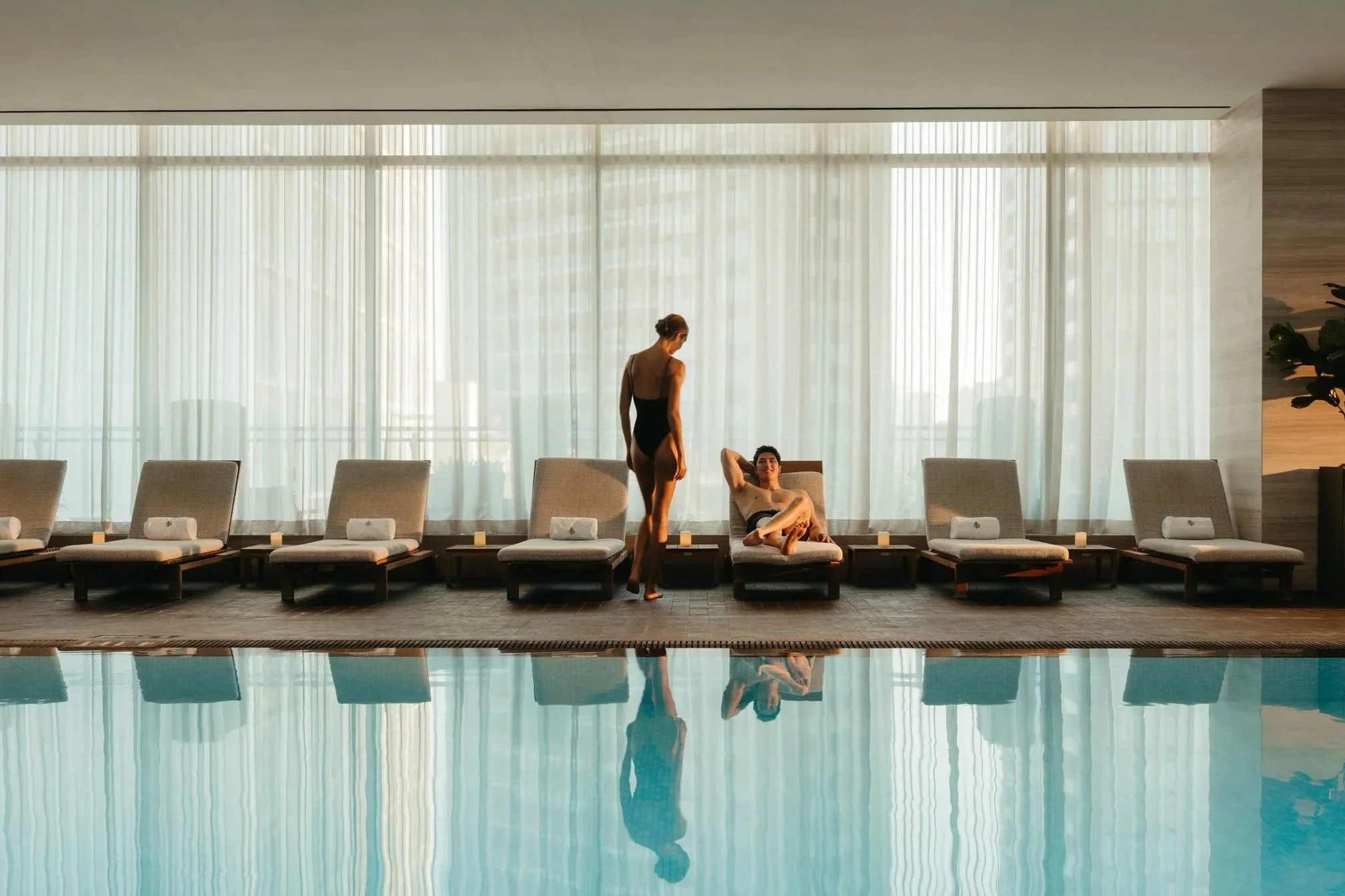 Indoor swimming pool area with a row of lounge chairs along the window. Two women in swimsuits are relaxing and conversing, one sitting and the other standing. The large windows with sheer curtains let in natural light, and the pool's reflection is v