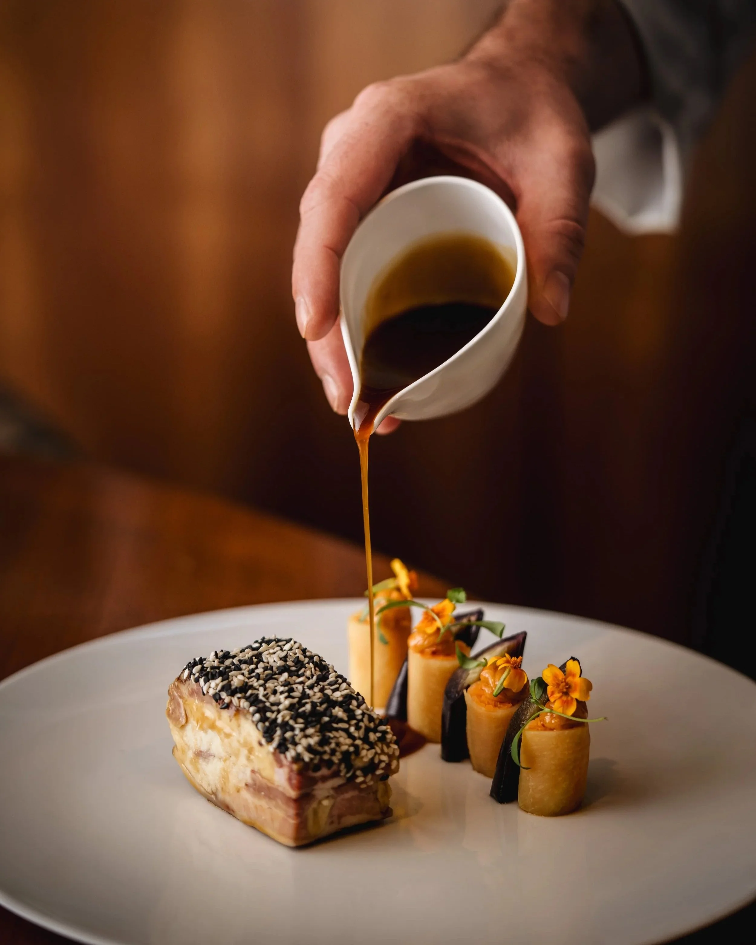 A close-up of a hand pouring dark sauce or gravy onto a gourmet dish with rolled food items and garnishes, on a white plate.