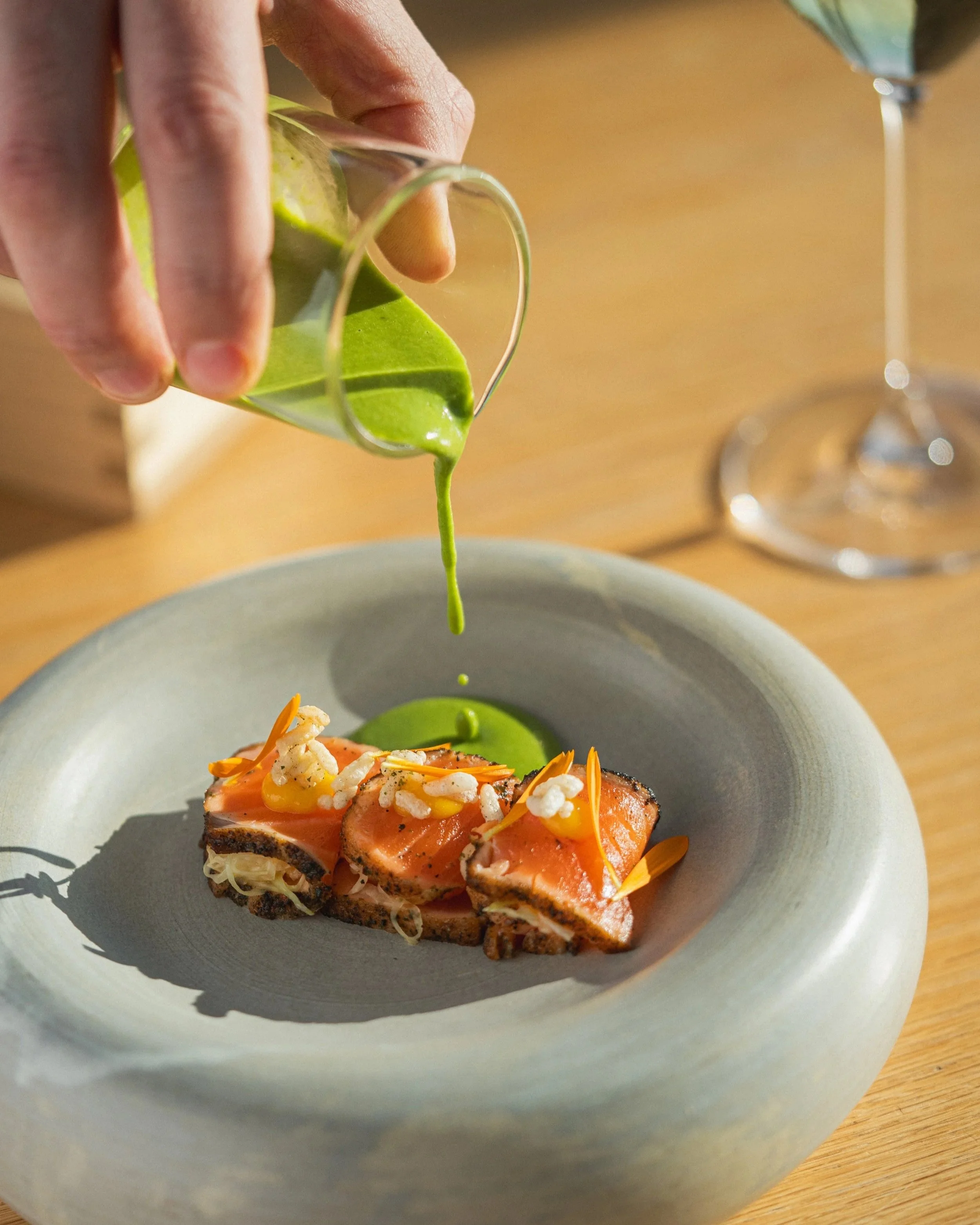 A close-up of a person pouring bright green sauce or oil from a small pitcher onto a piece of cooked salmon topped with shredded vegetables and garnishes, served on a grey plate.