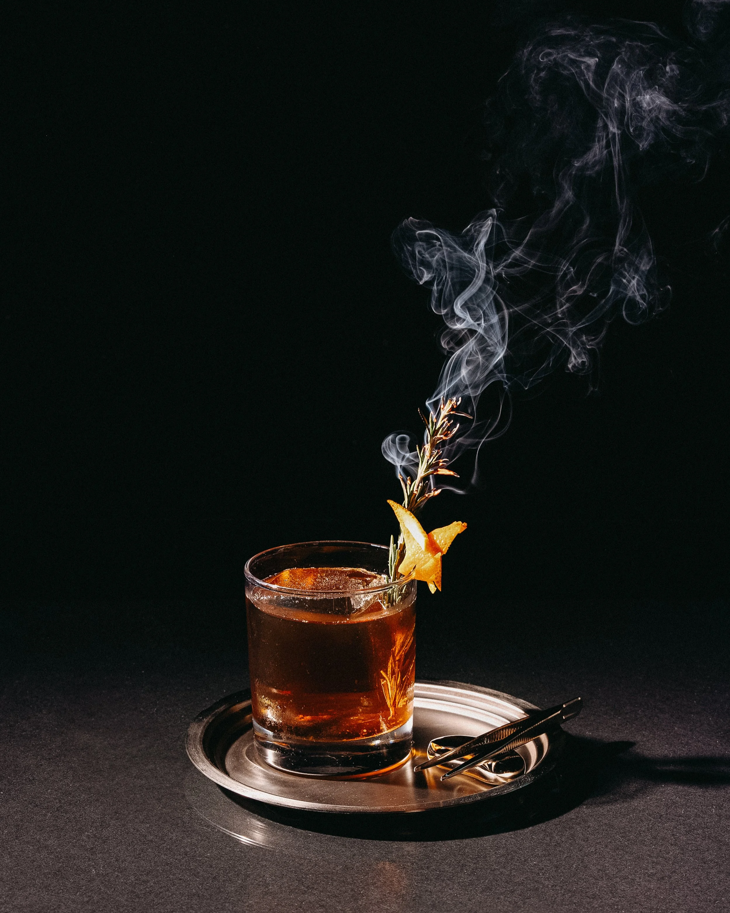 A glass of cold iced tea with a slice of lemon and a sprig of rosemary, placed on a metallic tray with ice tongs on a black surface, with wisps of steam or smoke rising from the drink.