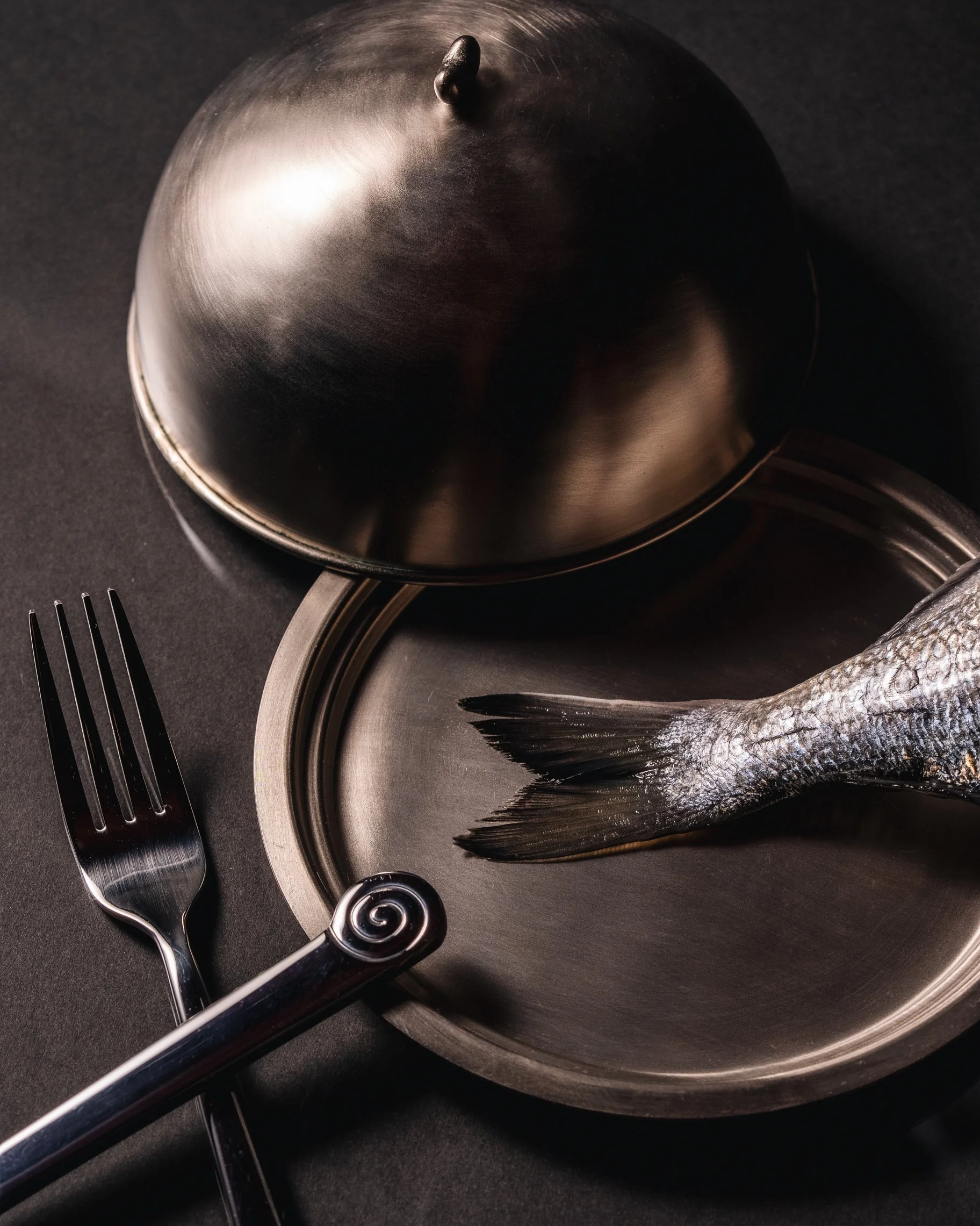 A fish lying on a metallic plate with a fork and a spoon nearby, all set on a dark surface.