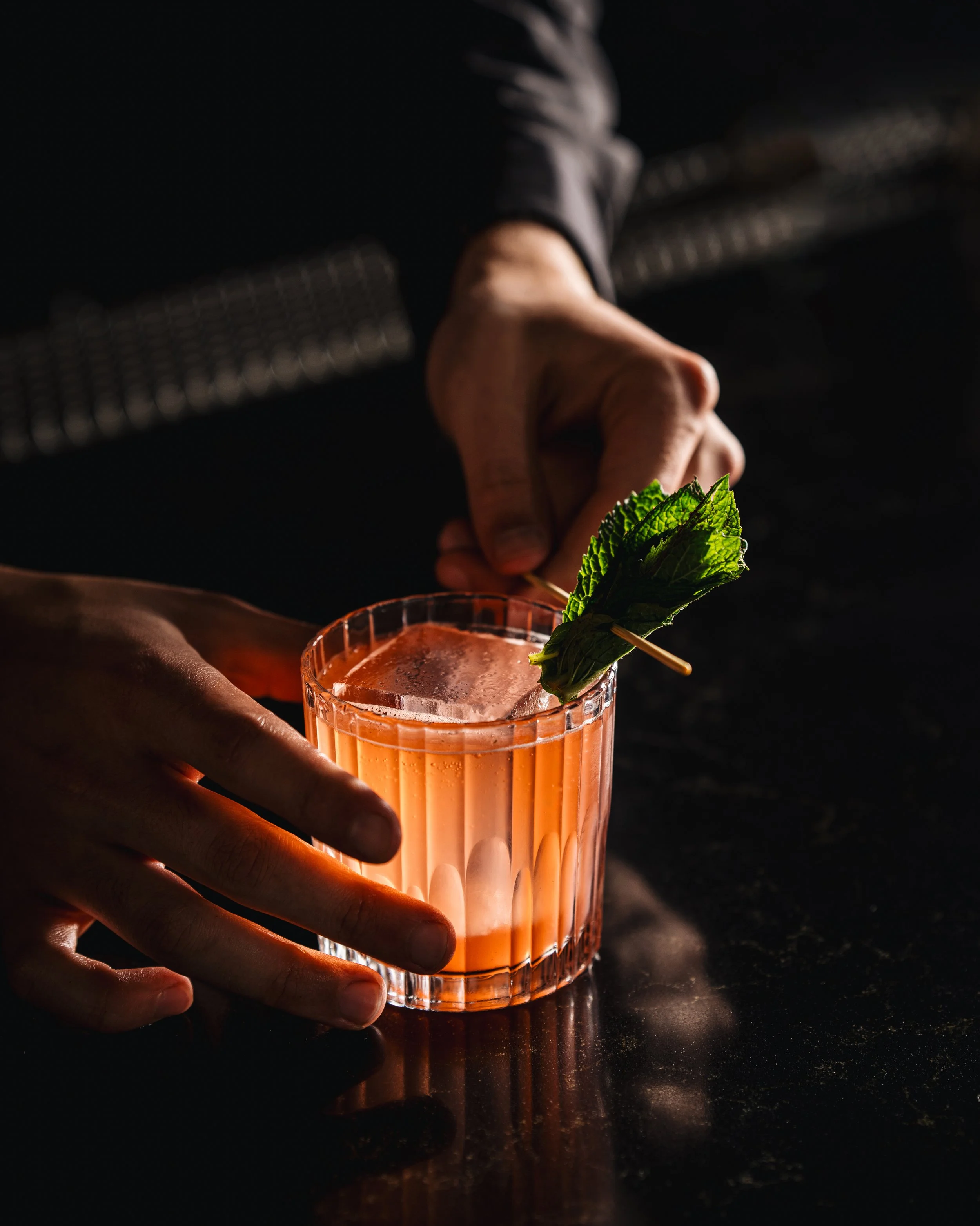 A hand holding a glass of pink cocktail garnished with a sprig of mint, with a dark background and a reflective surface.