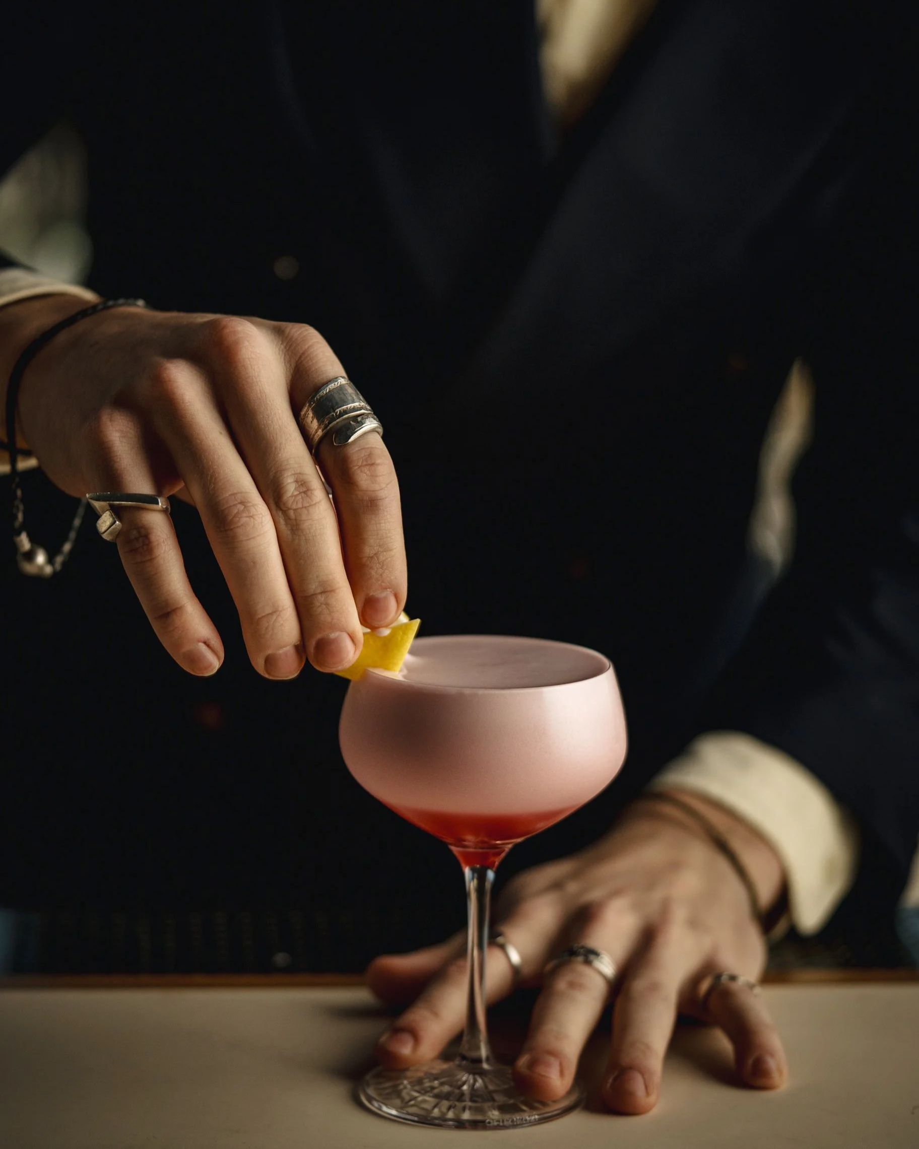 Close-up of a hand holding a lemon wedge above a pink cocktail glass, with fingers resting on a table, and a person wearing rings and bracelets in the background.