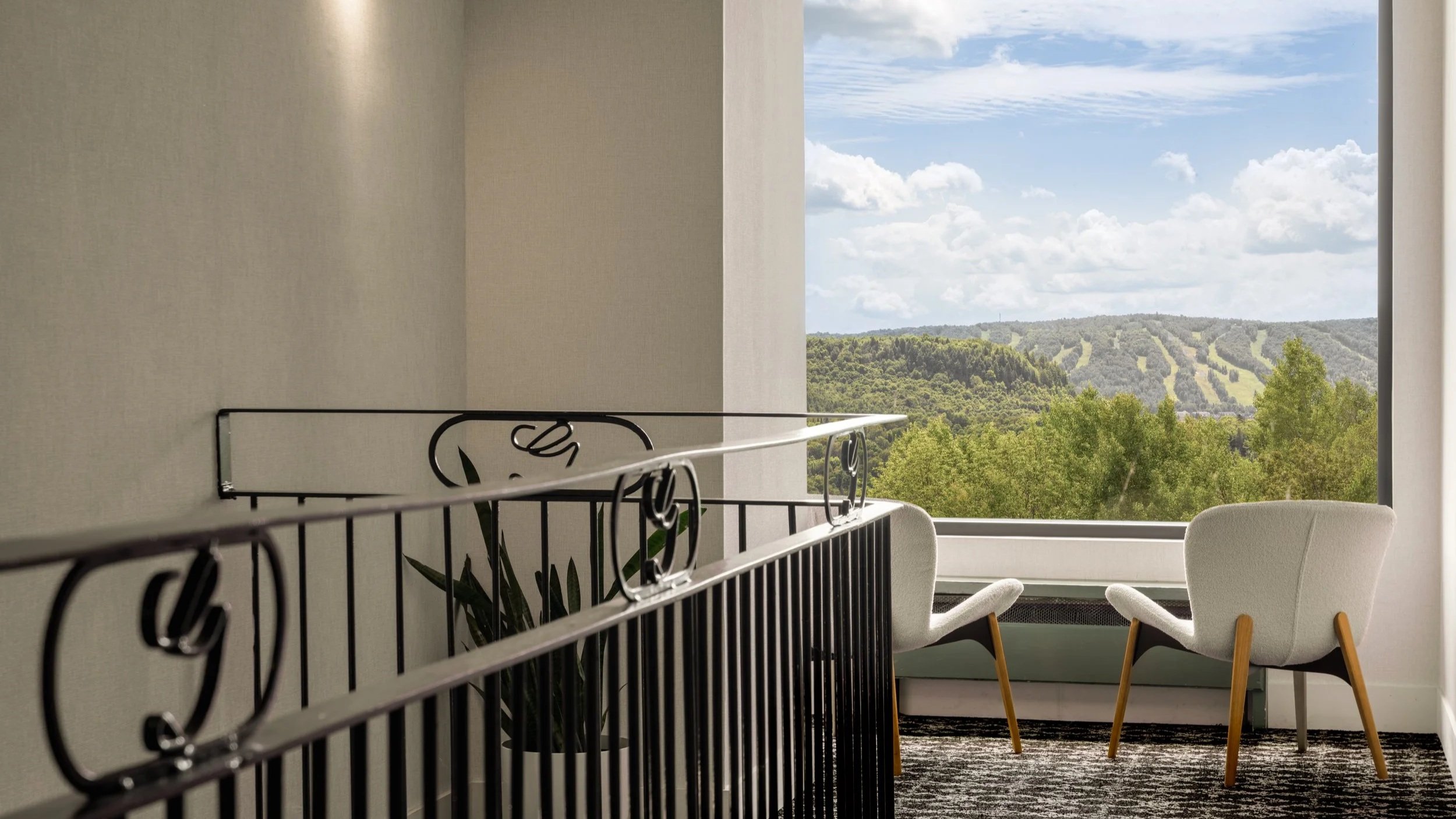 A cozy room with large window overlooking green hills and blue sky, two white armchairs, and a black metal railing in the foreground.