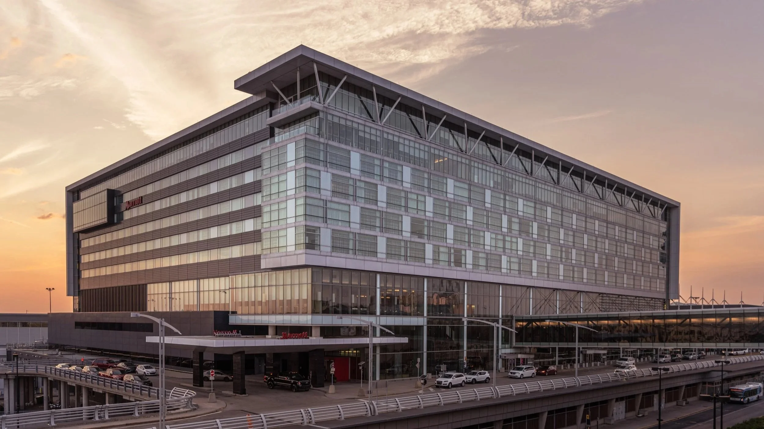 Modern multi-story hotel building with glass windows and a parking area in front, under a sunset sky.