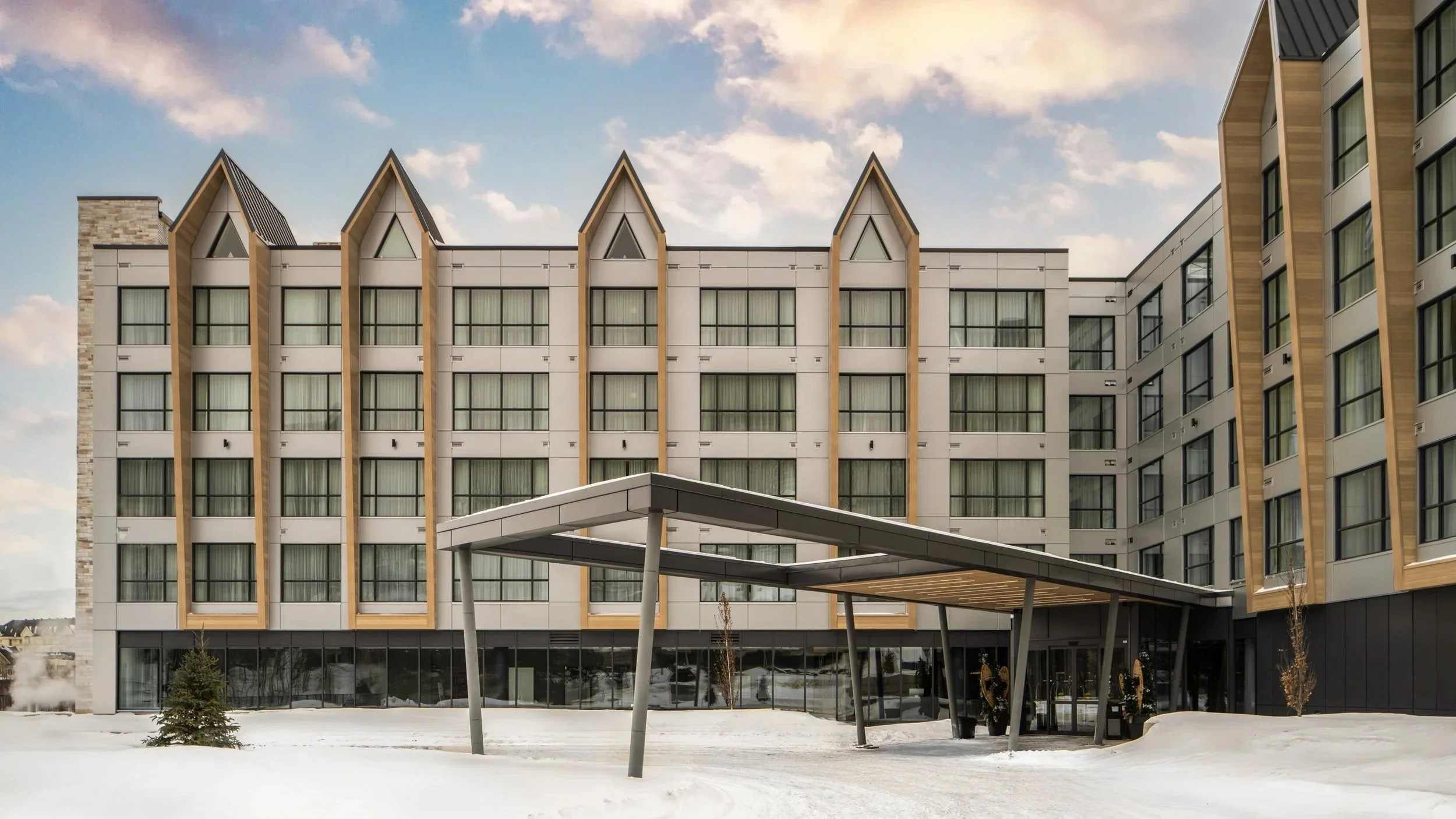 Modern multi-story building with large windows, wooden accents, and a covered entrance, set against a partly cloudy sky and snowy ground.