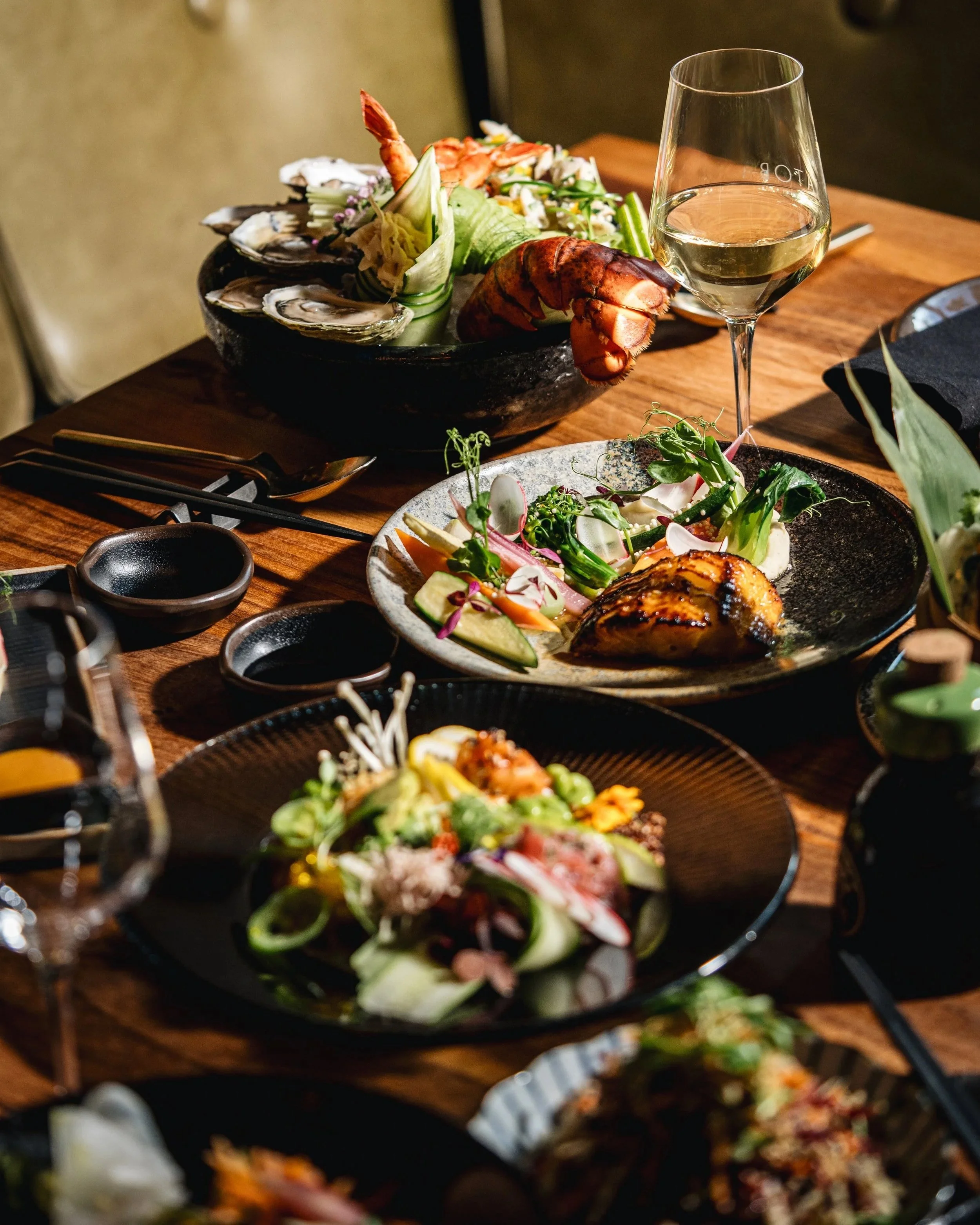 A table set with various dishes including oysters, lobster, salad, and a glass of white wine. The dishes are placed on a wooden table with black plates and utensils.